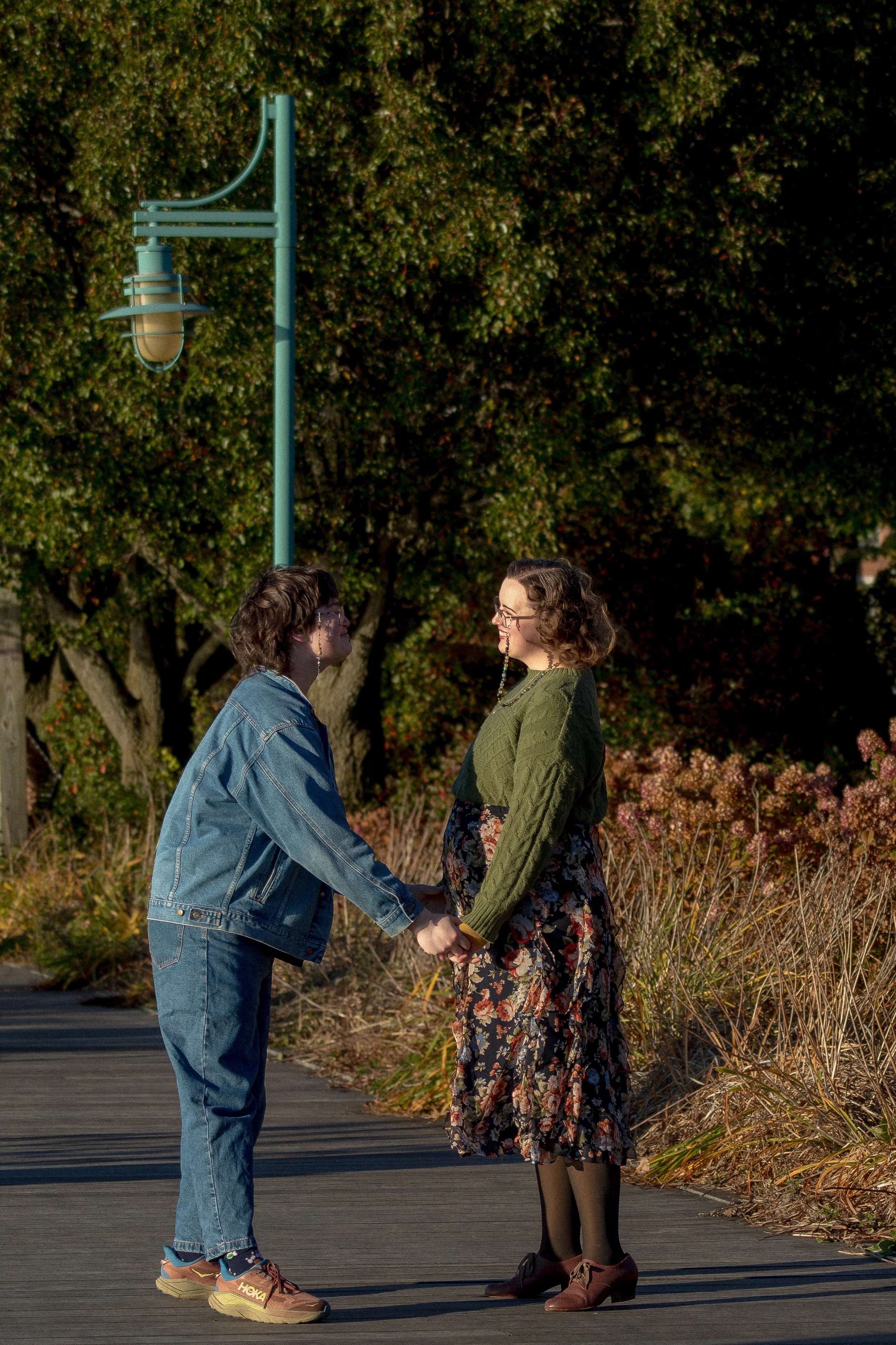 Two women holding hands and facing each other outdoors during daytime, surrounded by trees and plants, with a blue street lamp in the background.