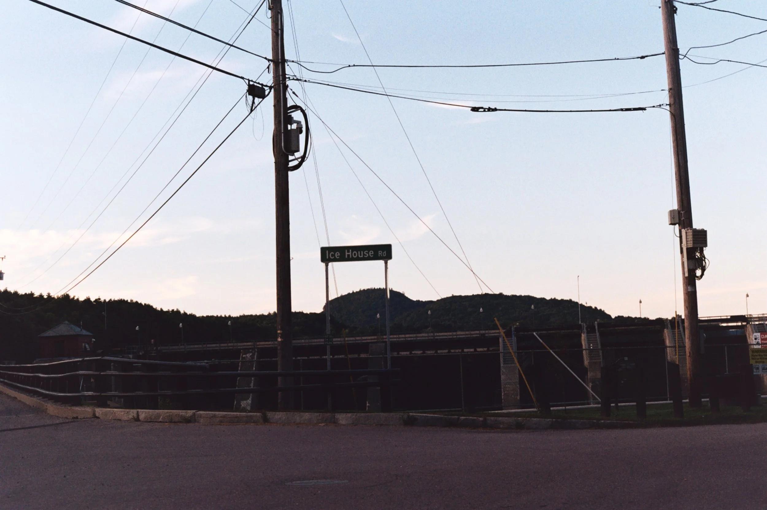 A rural scene with utility poles and power lines, a street sign reading 'Ice House Rd,' and mountains in the background during dusk or dawn.