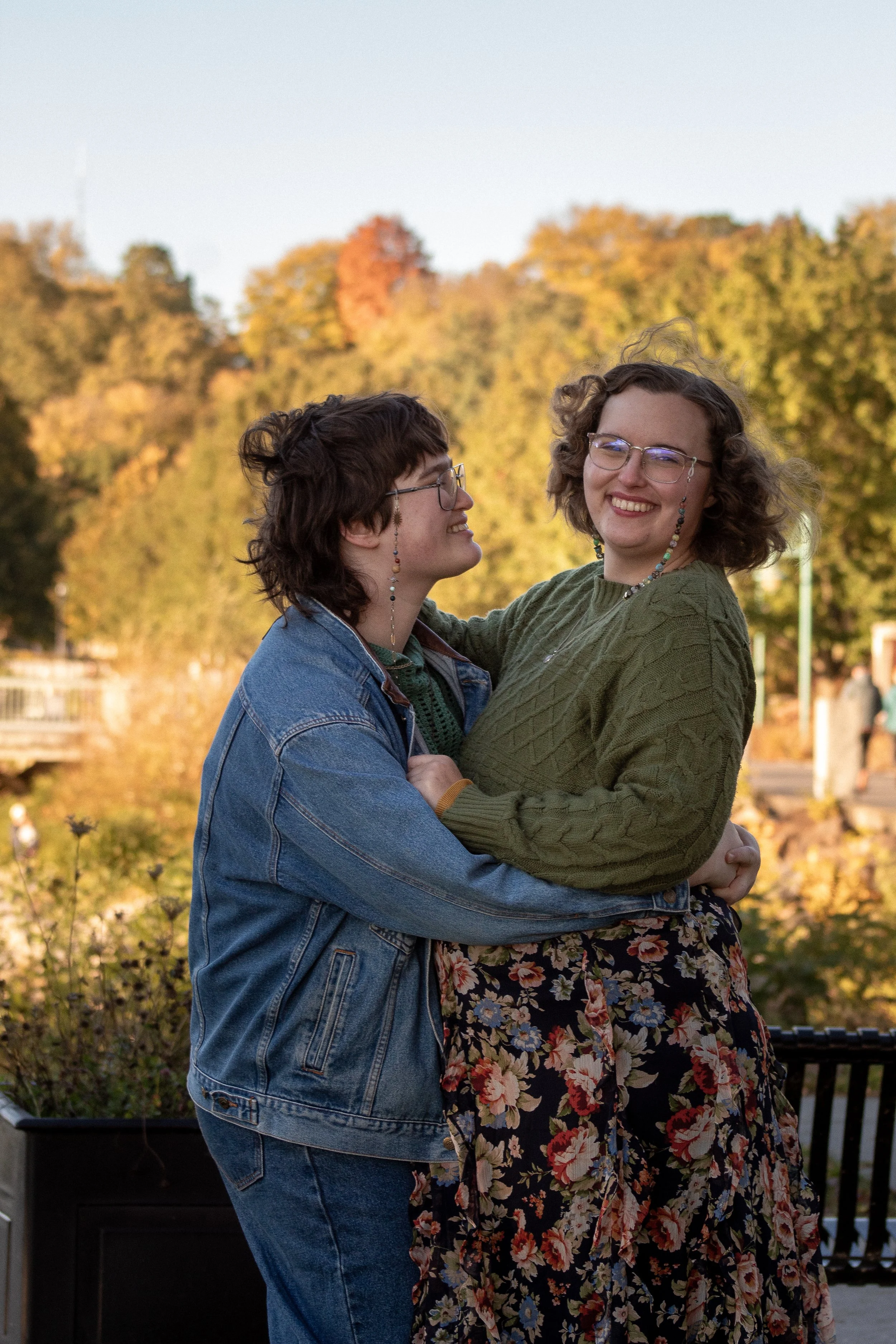 Two women hugging and smiling outdoors during autumn, with colorful fall trees in the background.