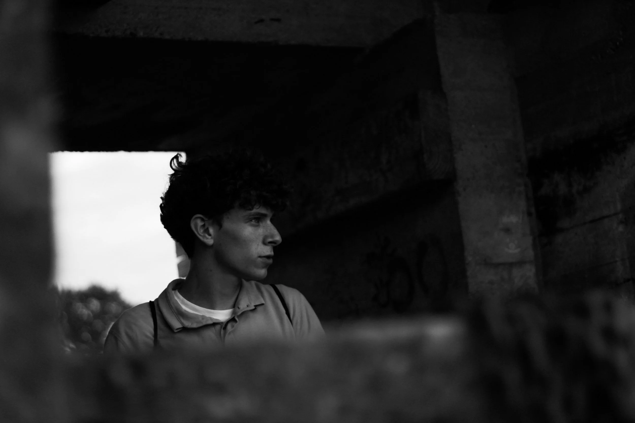 A black and white photo of a young man with curly hair, standing outdoors near a structure made of stone and wood, looking to his left.