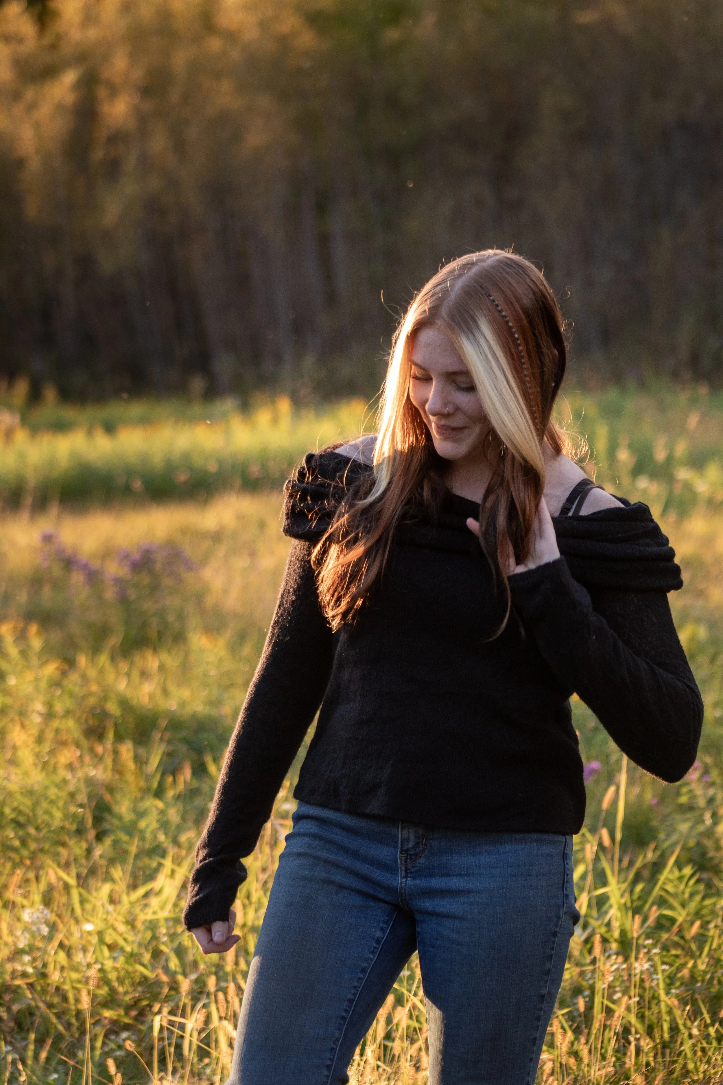 A woman with long hair, dressed in a black top and jeans, standing in a field during sunset, with her eyes closed and a peaceful expression.