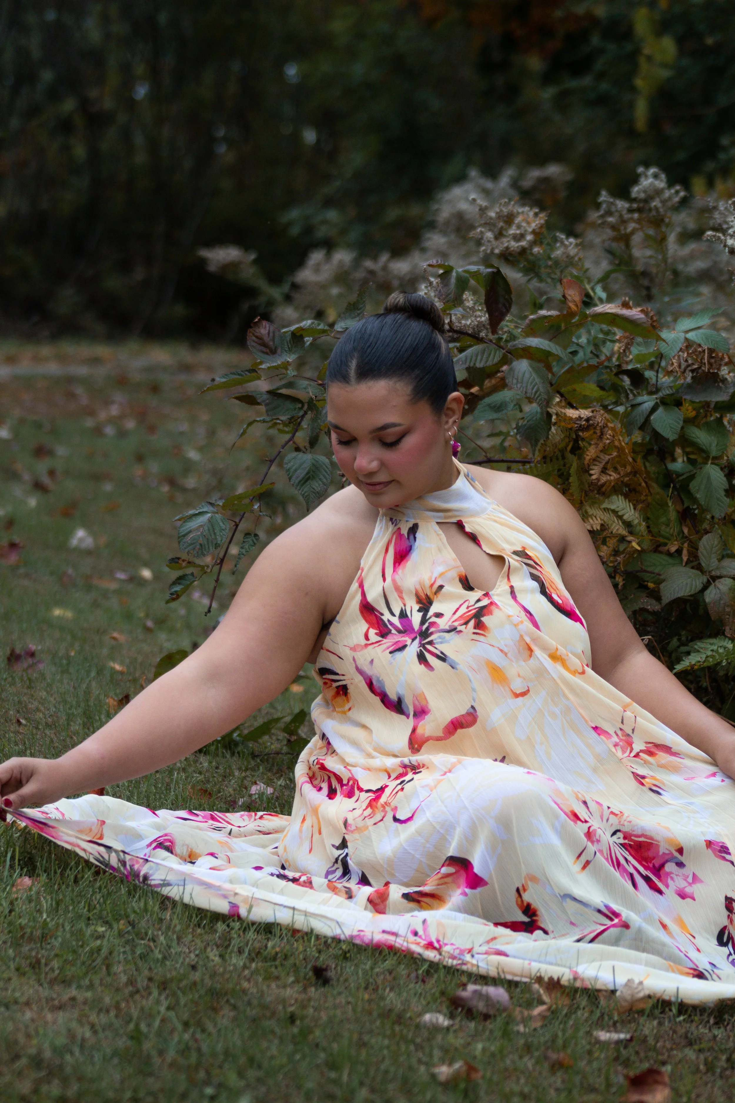 A woman with dark hair styled in a bun, wearing a colorful floral dress, sitting on the grass outdoors, surrounded by plants and trees, looking down with a peaceful expression.