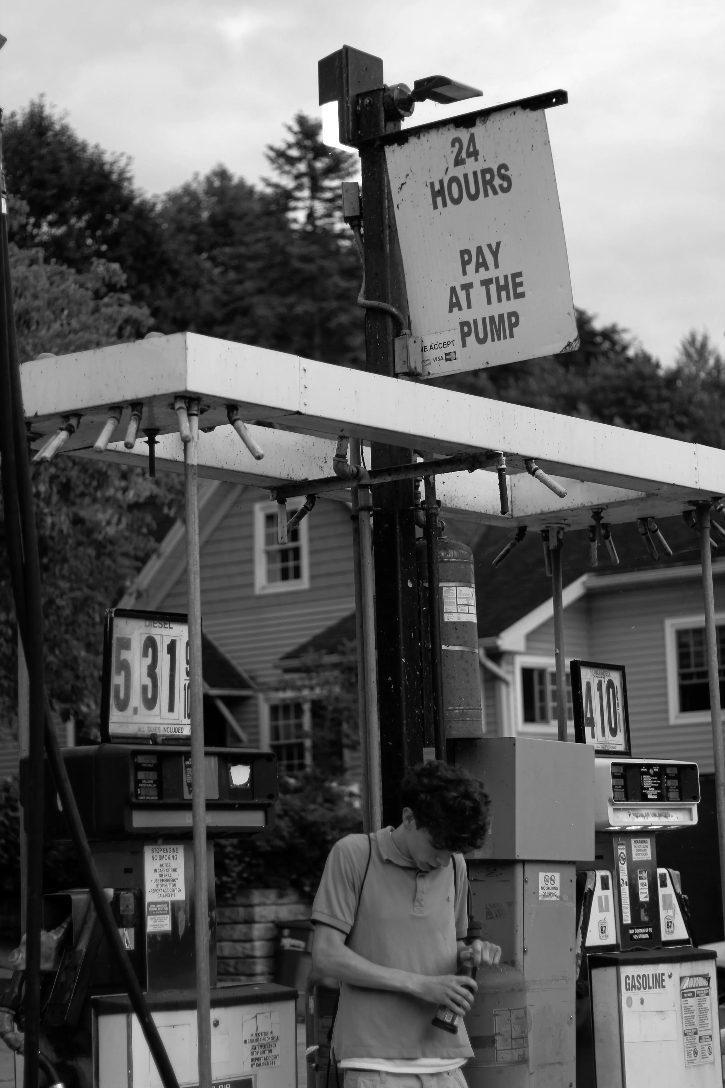 A young man changing the oil in his car at a gas station, with gasoline pumps and a gas station sign in the background.