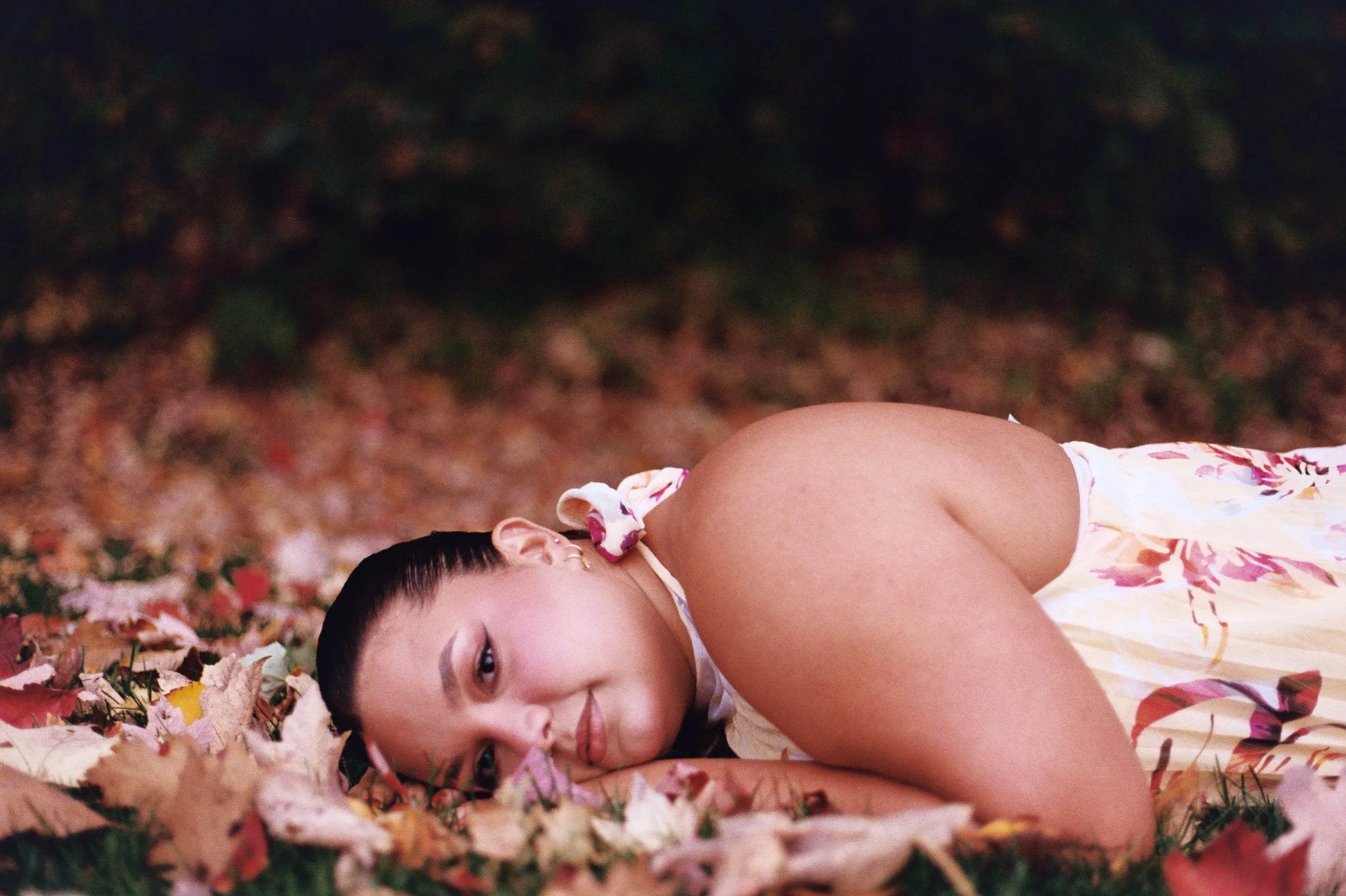A young woman lying on the ground covered with fallen autumn leaves, with her eyes open and looking at the camera, wearing a floral top and earrings.