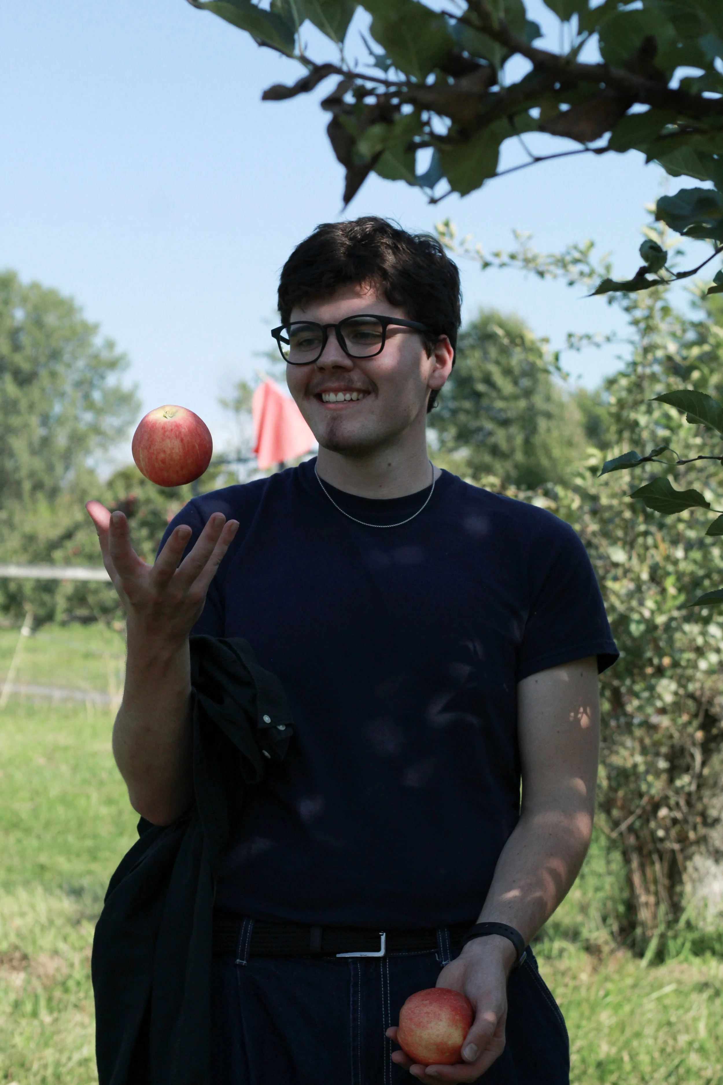 A young man wearing glasses and a navy blue t-shirt is smiling while balancing an apple on his finger and holding another apple in his hand outdoors near trees and a grassy area.