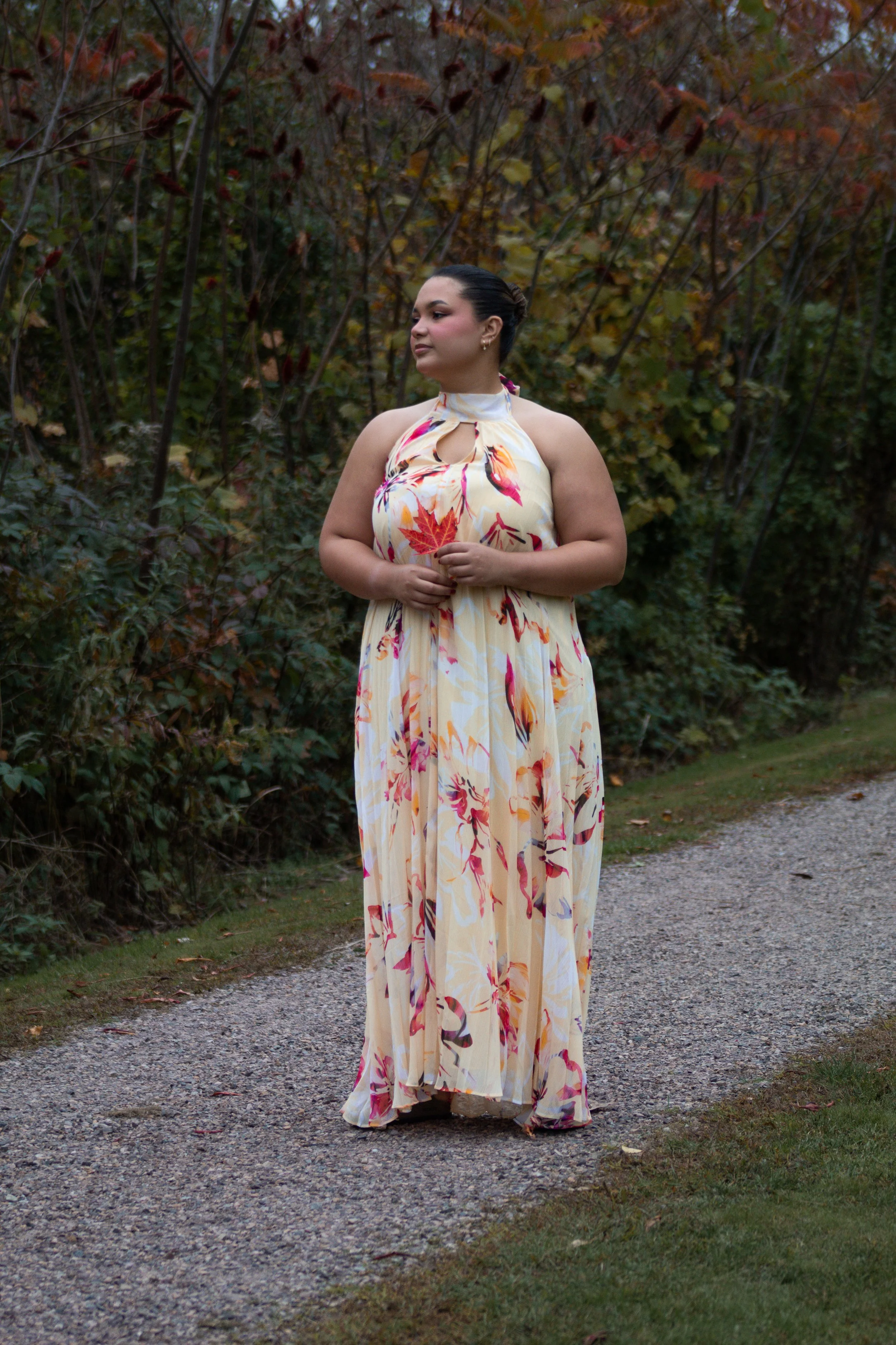 A woman in a sleeveless, floral maxi dress standing on a gravel pathway surrounded by autumn foliage.