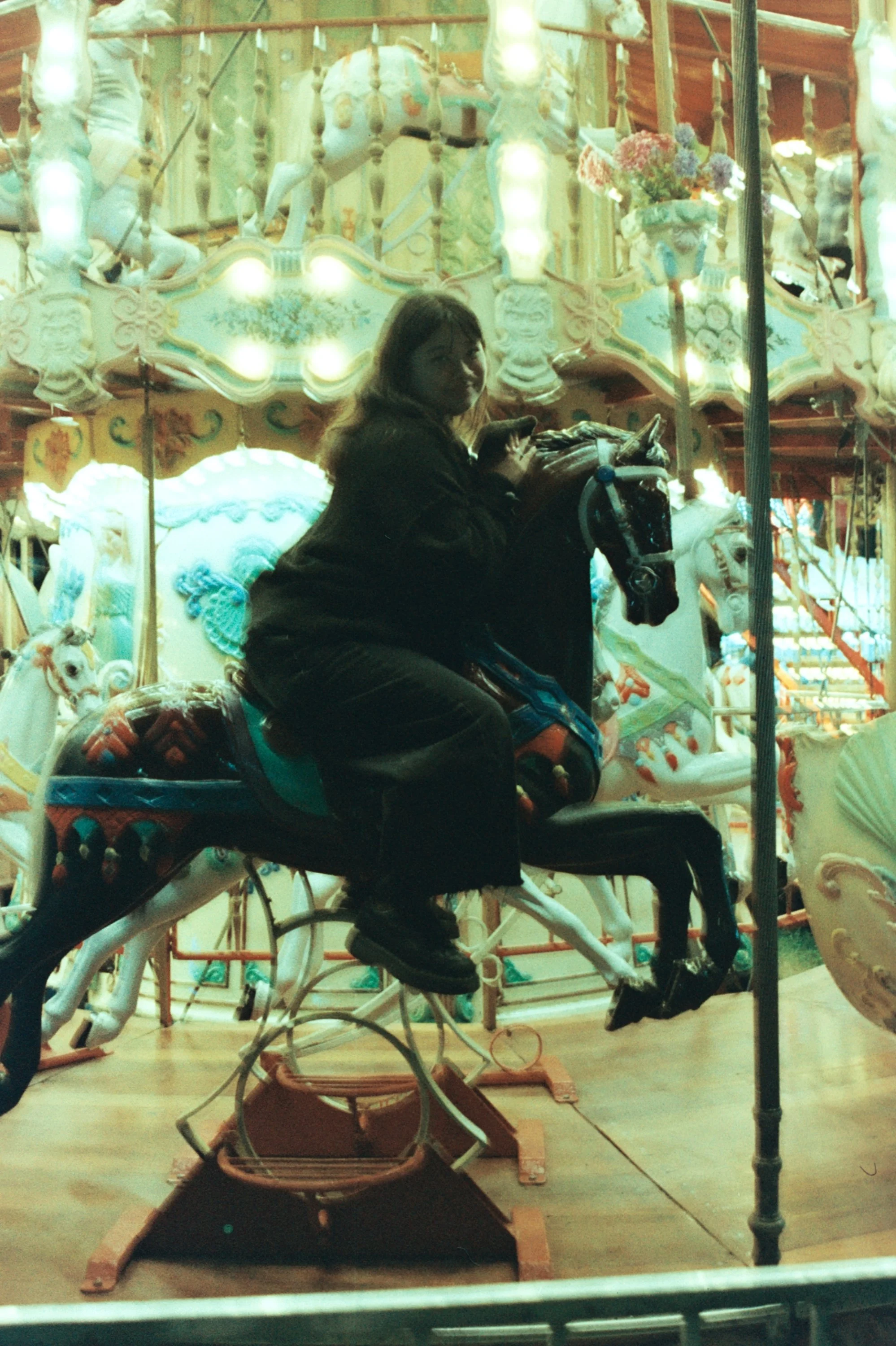 A woman sitting on a carousel horse inside an amusement park, with decorative lights and floral arrangements in the background.