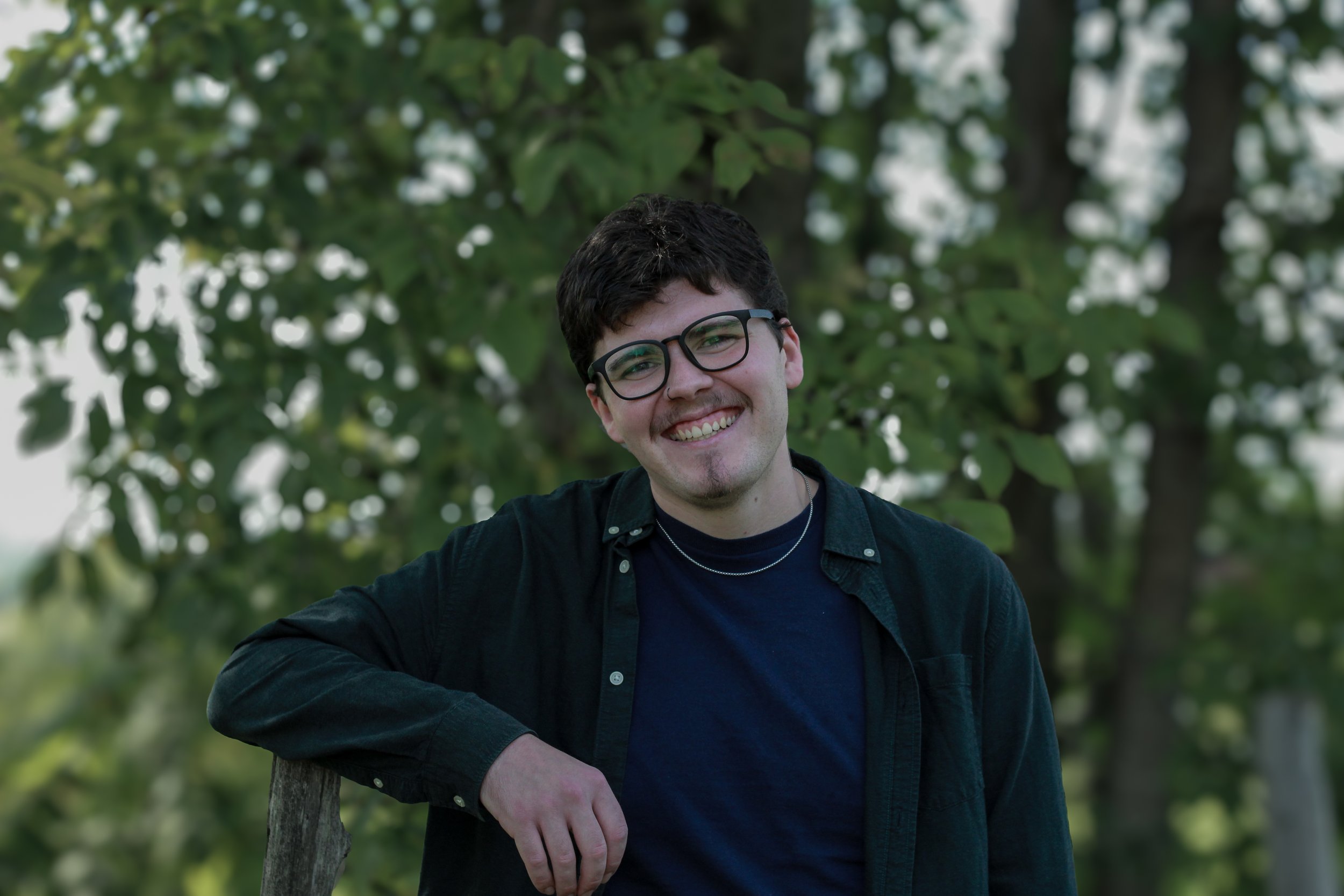 A smiling young man with black glasses and dark hair, wearing a black jacket over a navy blue shirt, standing outdoors with green trees in the background.