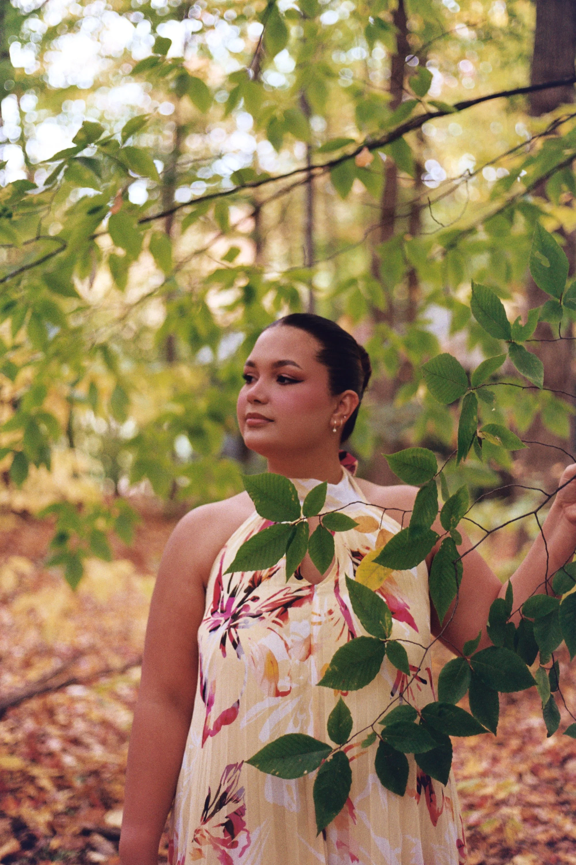 A woman with dark hair styled in an elegant updo, wearing earrings, in a forest setting with green and yellow foliage, wearing a colorful floral dress.