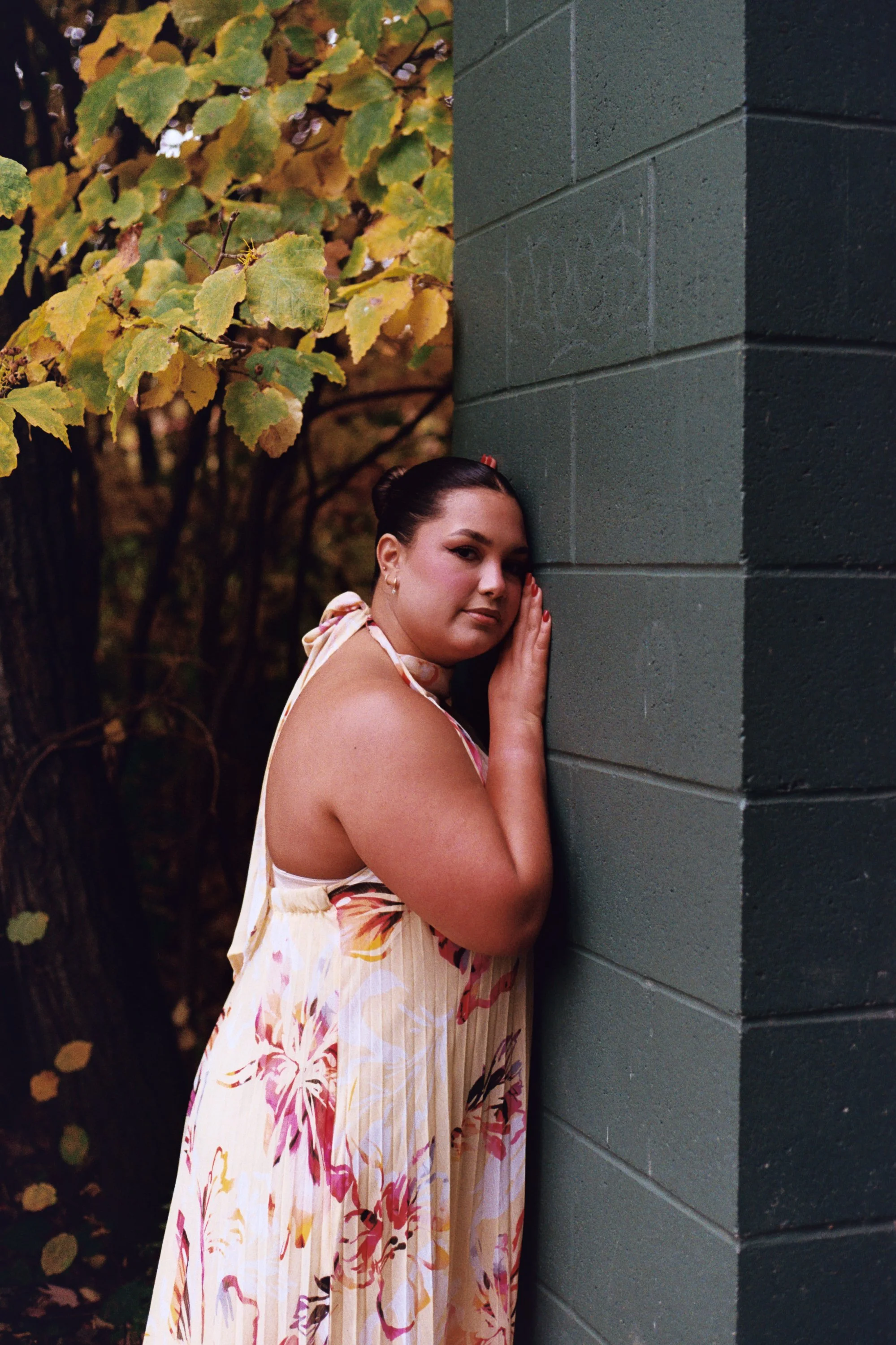 A woman with dark hair tied back, wearing a sleeveless floral dress, is leaning against a gray brick wall with her hand on it, outdoors during fall, with yellow and green leaves in the background.