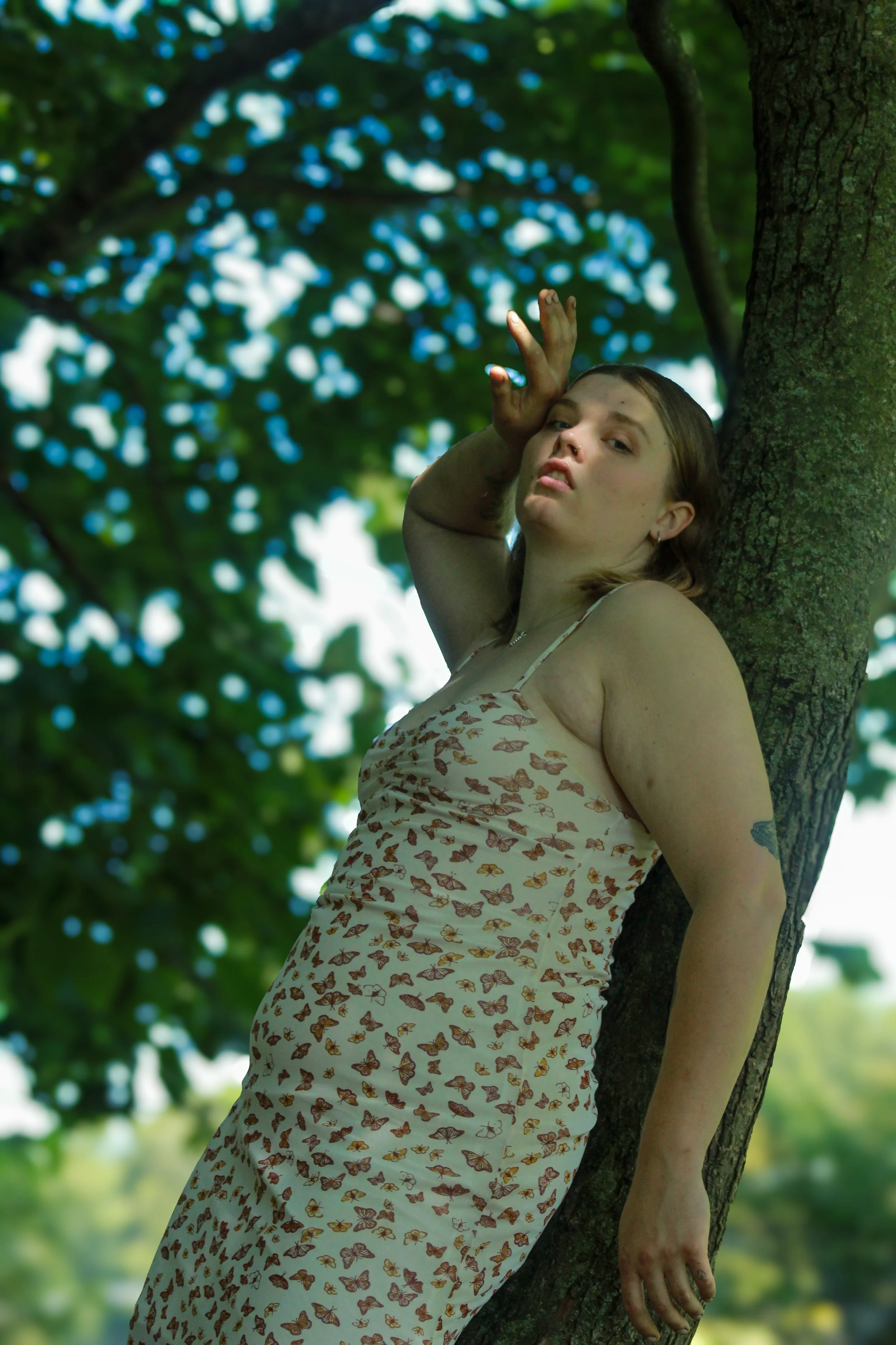 A young woman with a septum piercing and wearing a butterfly-patterned dress leans against a tree outdoors, surrounded by blurry green leaves and light.