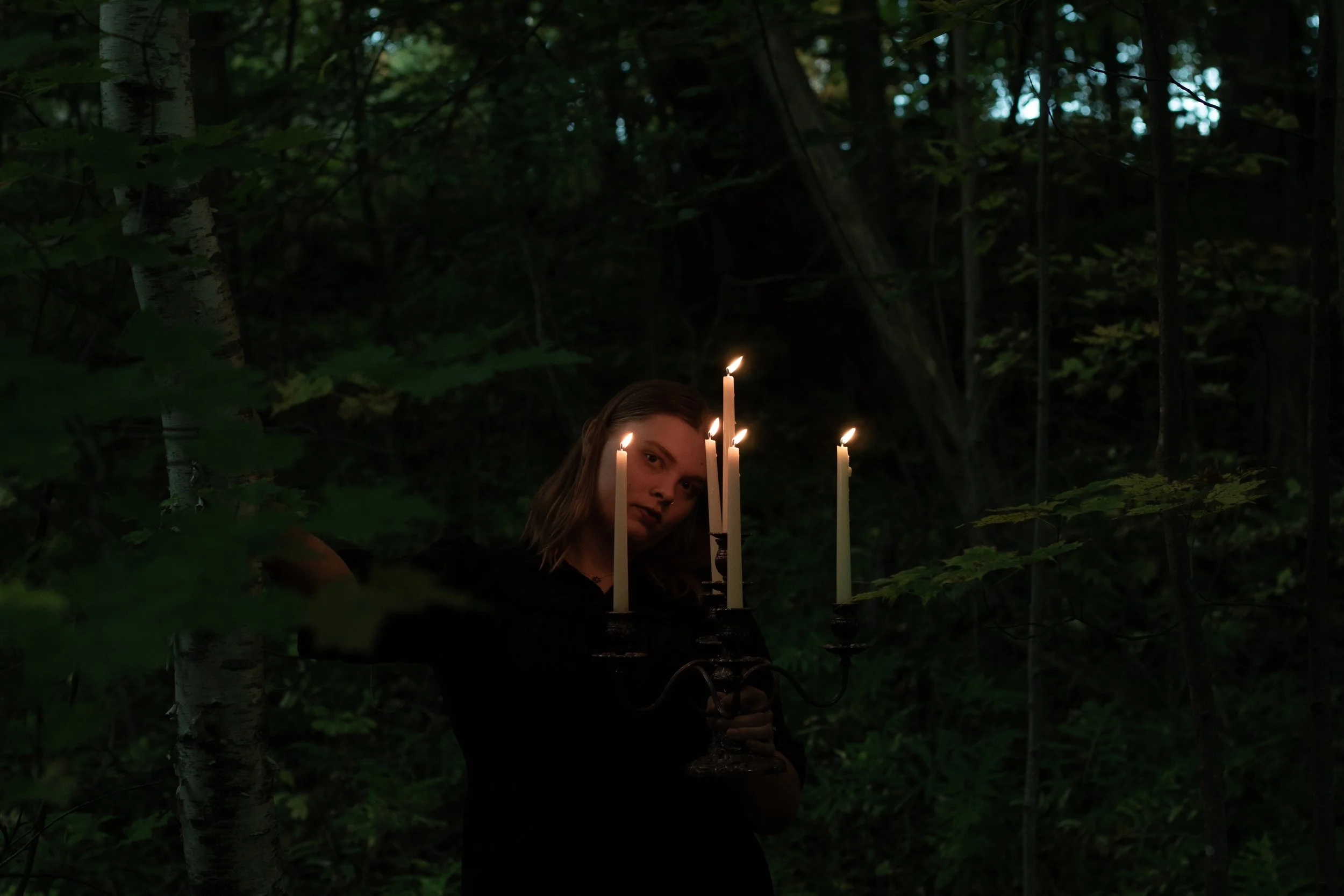 Young woman with long brown hair holding candles in a dark forest at night.