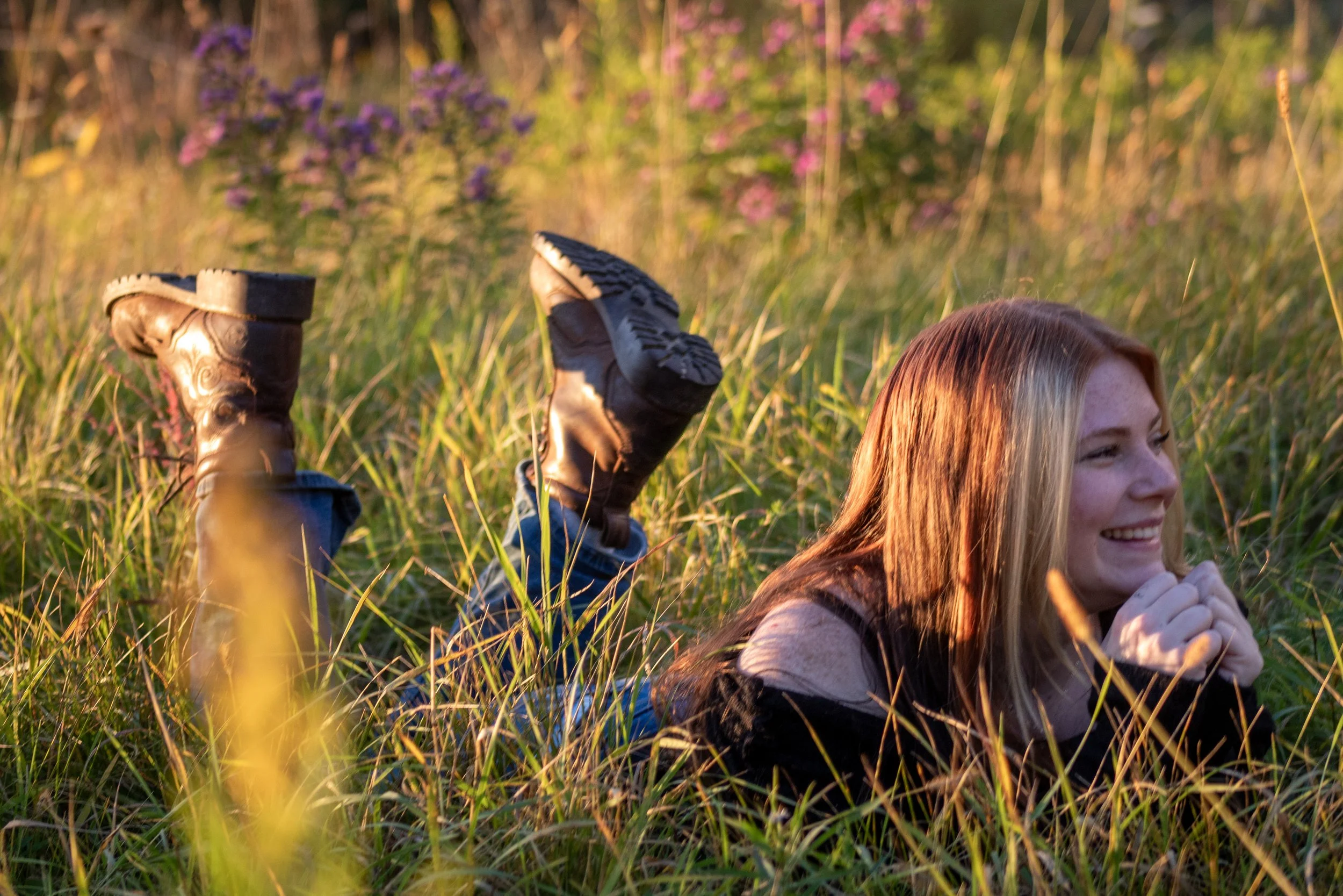 A girl with long reddish hair sitting in tall grass, smiling, with her hands clasped together. In the background, a person is lying on their back with their legs in the air, wearing boots and jeans, in a field with pink and purple flowers, during sun