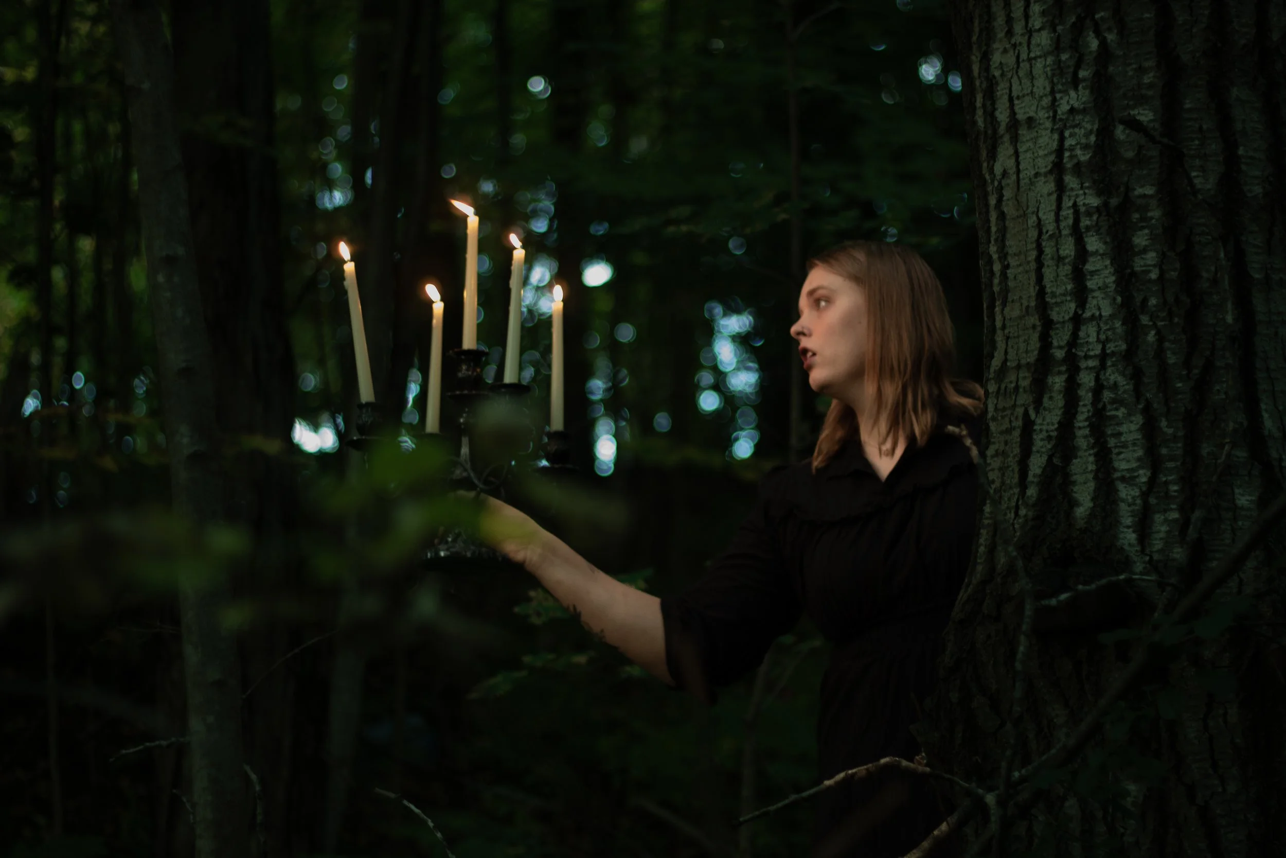 A young woman in dark clothing holding a candelabrum with lit candles in a dense, dark forest at night.