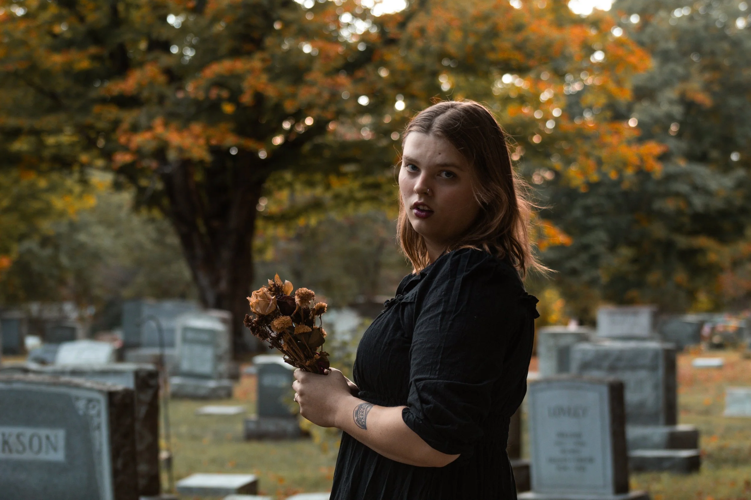 A young woman with red hair and a septum piercing holding a bouquet of dried flowers in a cemetery with autumn-colored trees in the background.