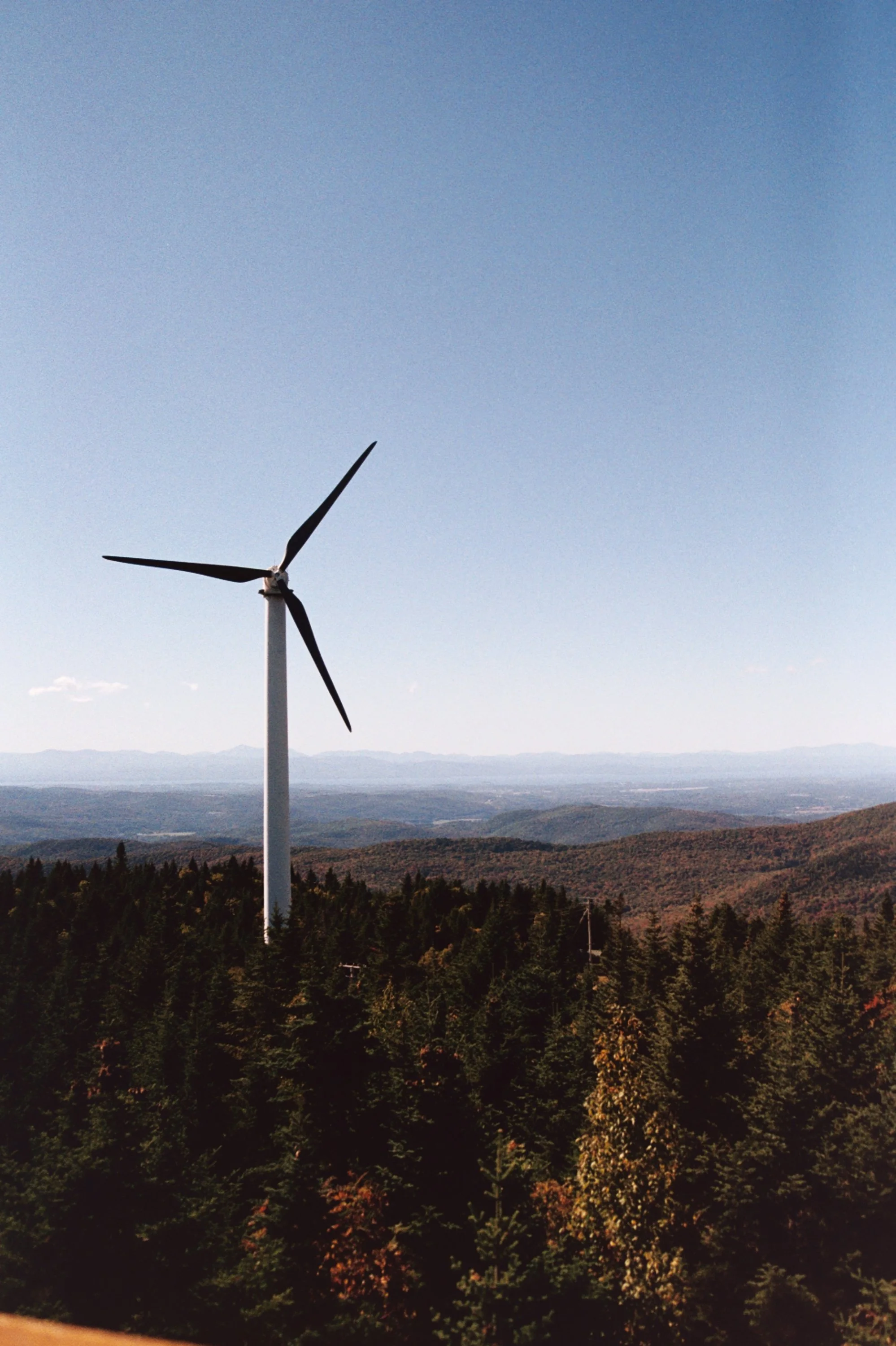 A large wind turbine with three blades standing in a landscape of trees and mountains under a clear blue sky.