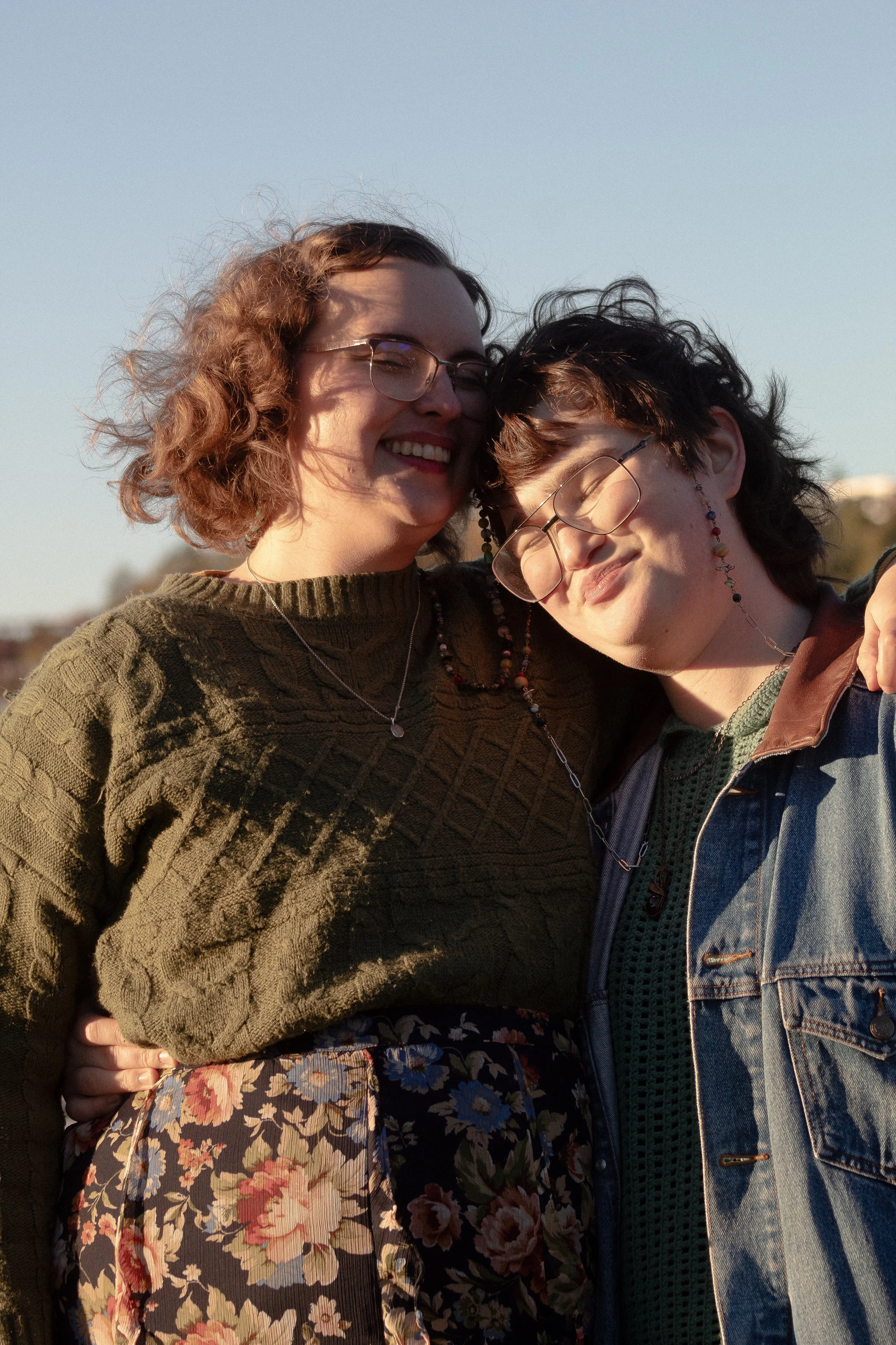 Two women smiling and hugging outdoors during sunset, one wearing a green sweater and floral skirt, the other in a denim jacket and glasses.