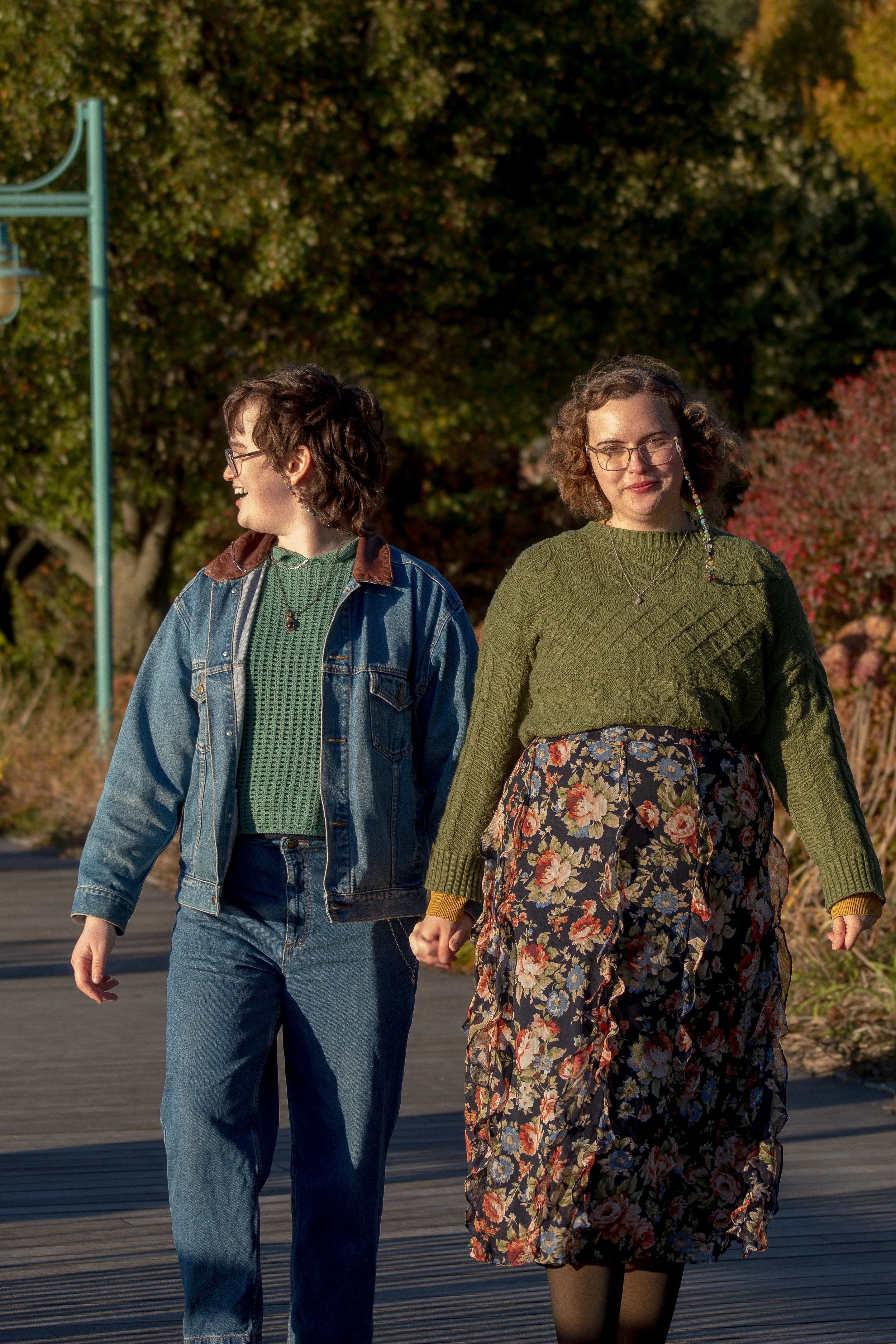 Two young women walking outdoors on a sunny day, surrounded by autumn foliage. One woman has short brown hair, glasses, and is wearing a denim jacket over a green sweater. The other woman has curly brown hair, glasses, and is wearing a green sweater 