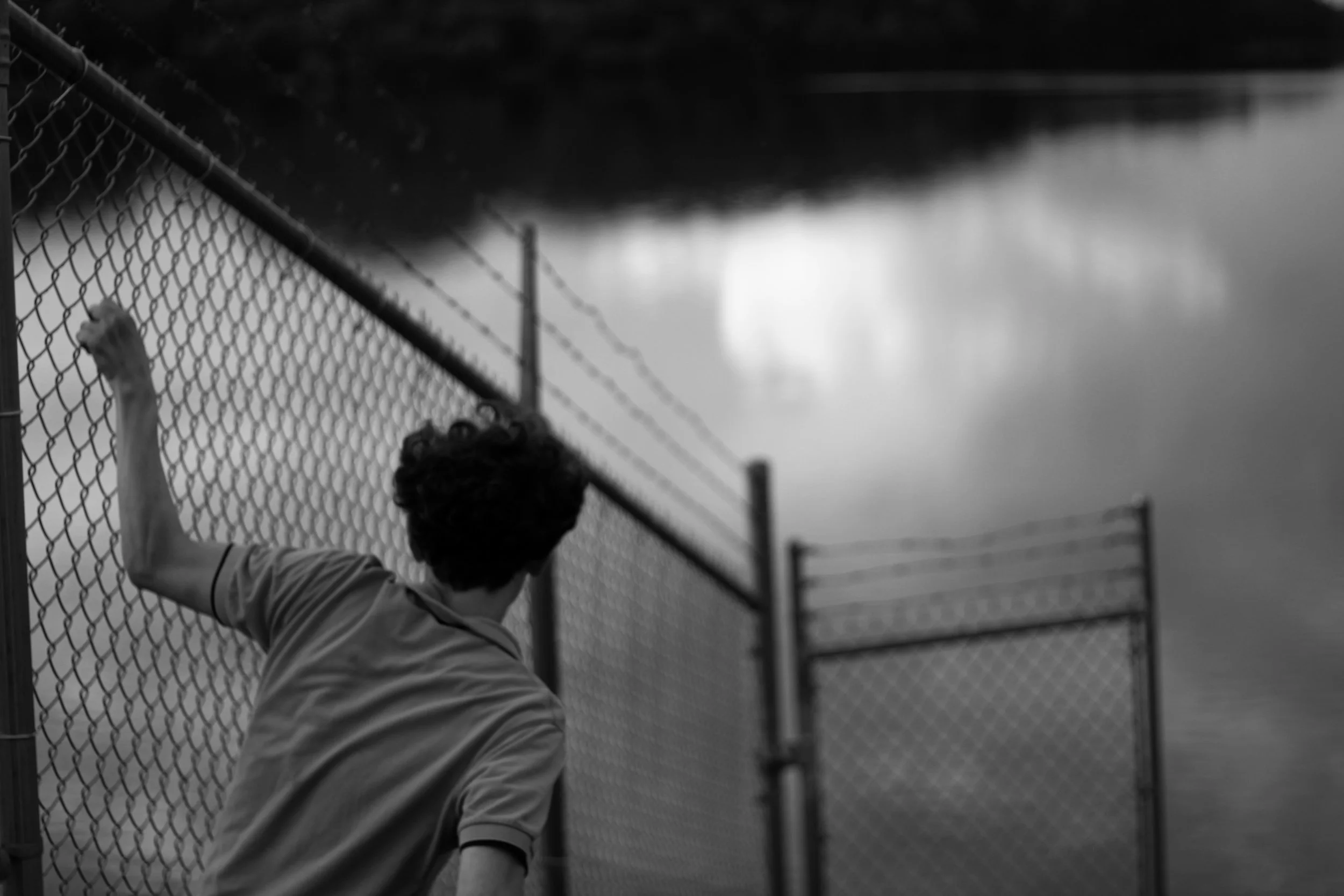 A person with curly hair and a striped shirt is leaning on a chain-link fence by a body of water, with trees and the sky reflected in the water.