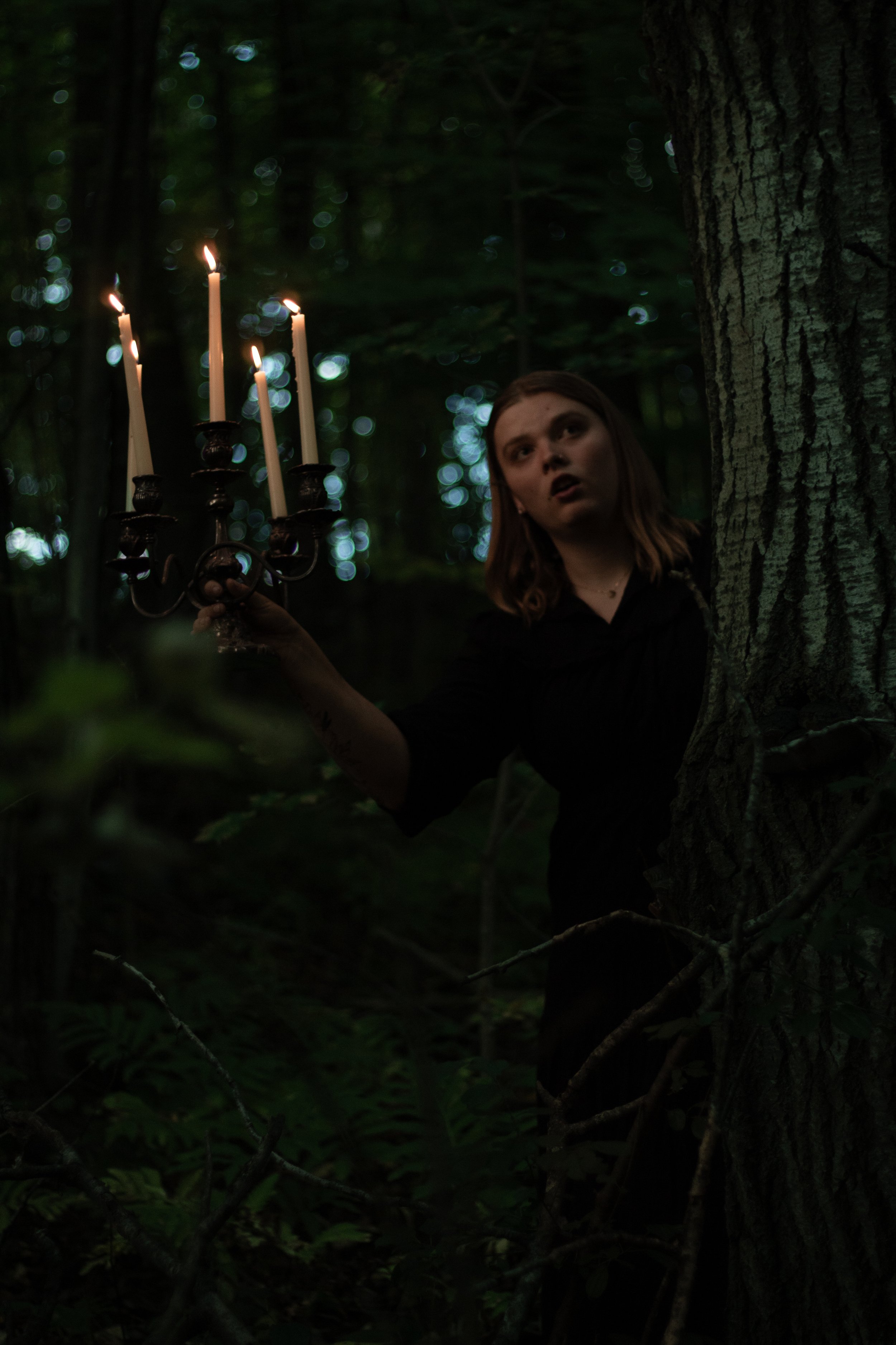 A woman in a dark forest holding a black candelabrum with lit candles, with a surprised or shocked expression on her face, partially obscured by a tree.