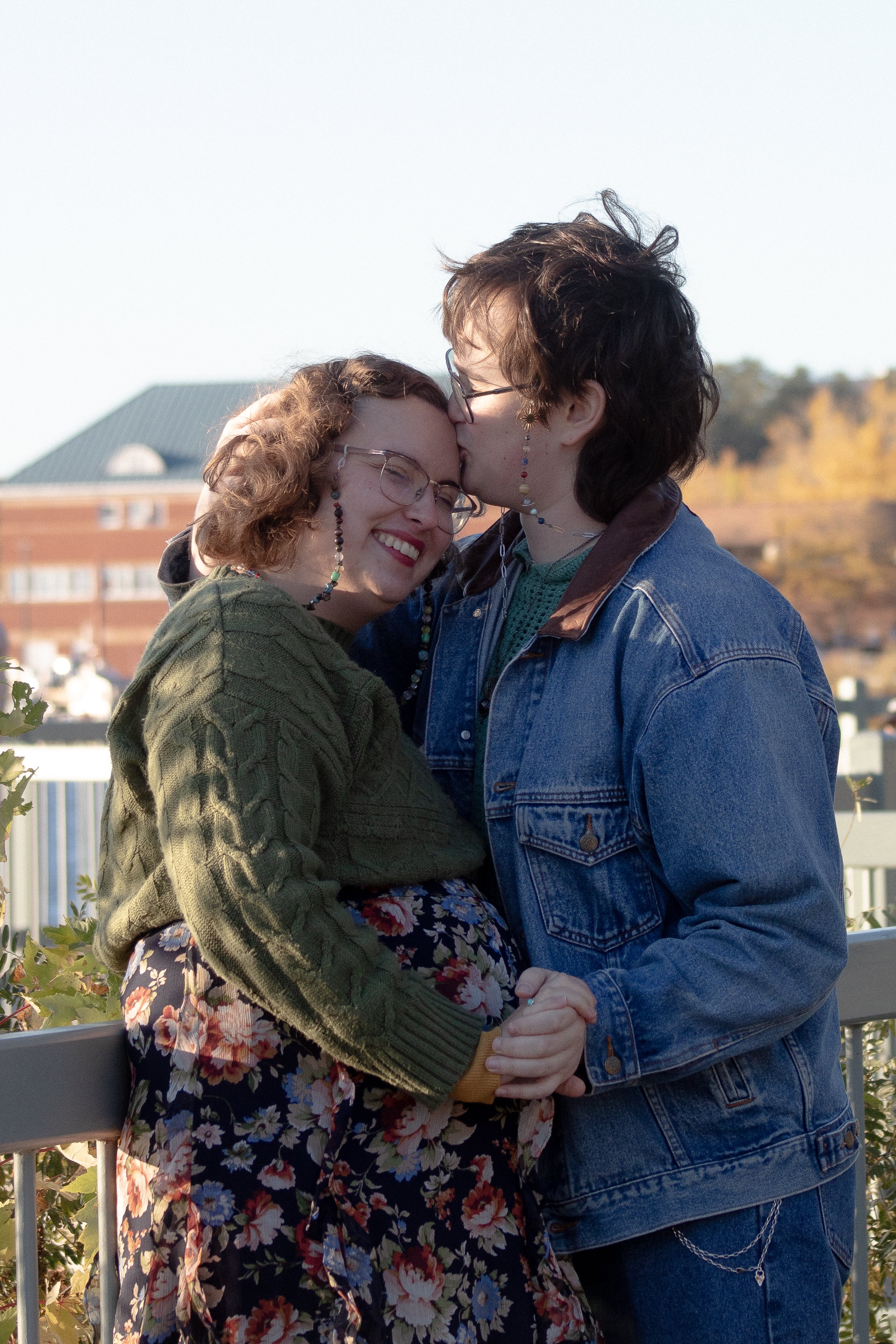 A woman with curly hair and glasses wearing a green sweater and floral skirt, smiling as she holds hands with a man with short dark hair, glasses, and earrings, wearing a denim jacket as he kisses her on the forehead. They are outdoors with a buildin