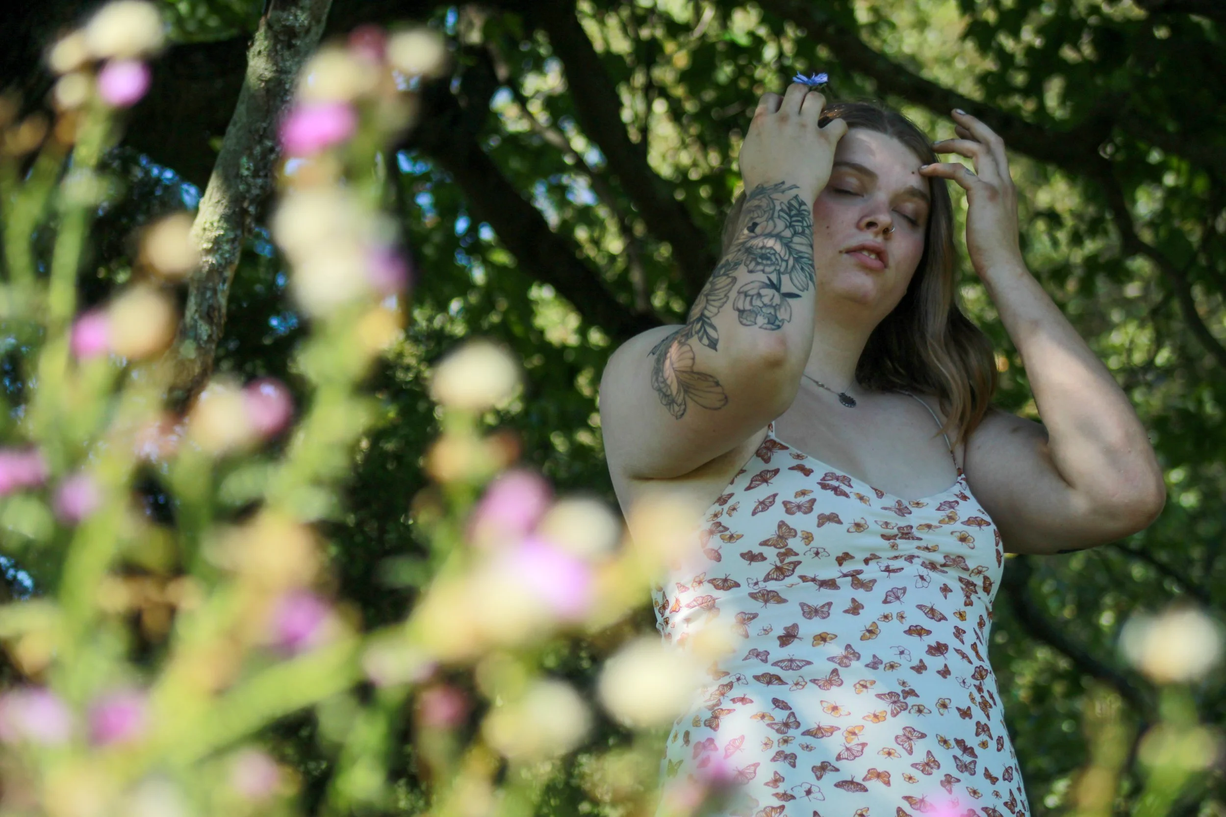 A woman with tattoos on her arms, wearing a dress with butterfly print, standing outdoors under green trees, touching her hair with both hands, with blurry pink flowers in the foreground.