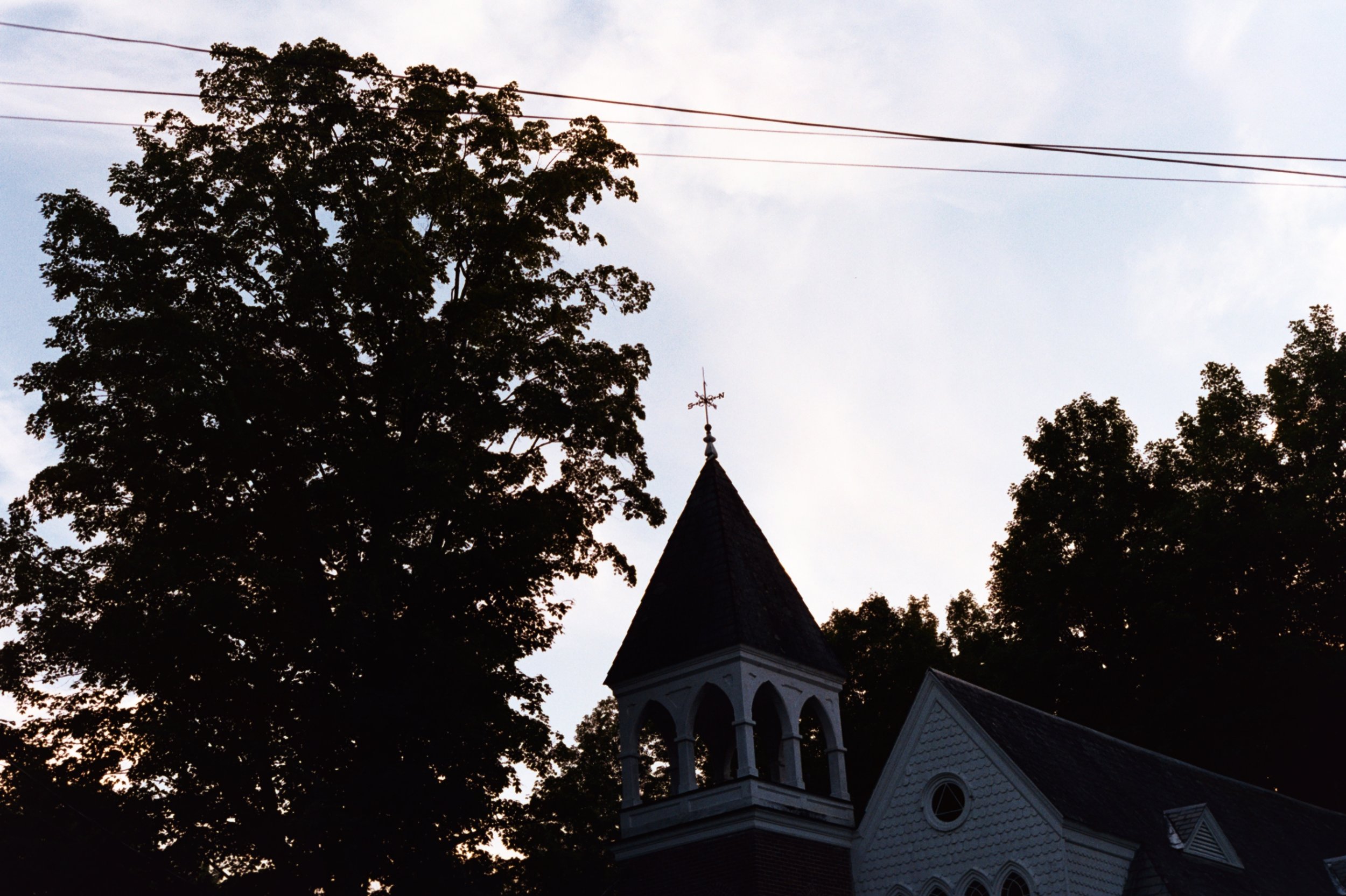 A church with a tall steeple, surrounded by trees, under a cloudy sky with power lines overhead.