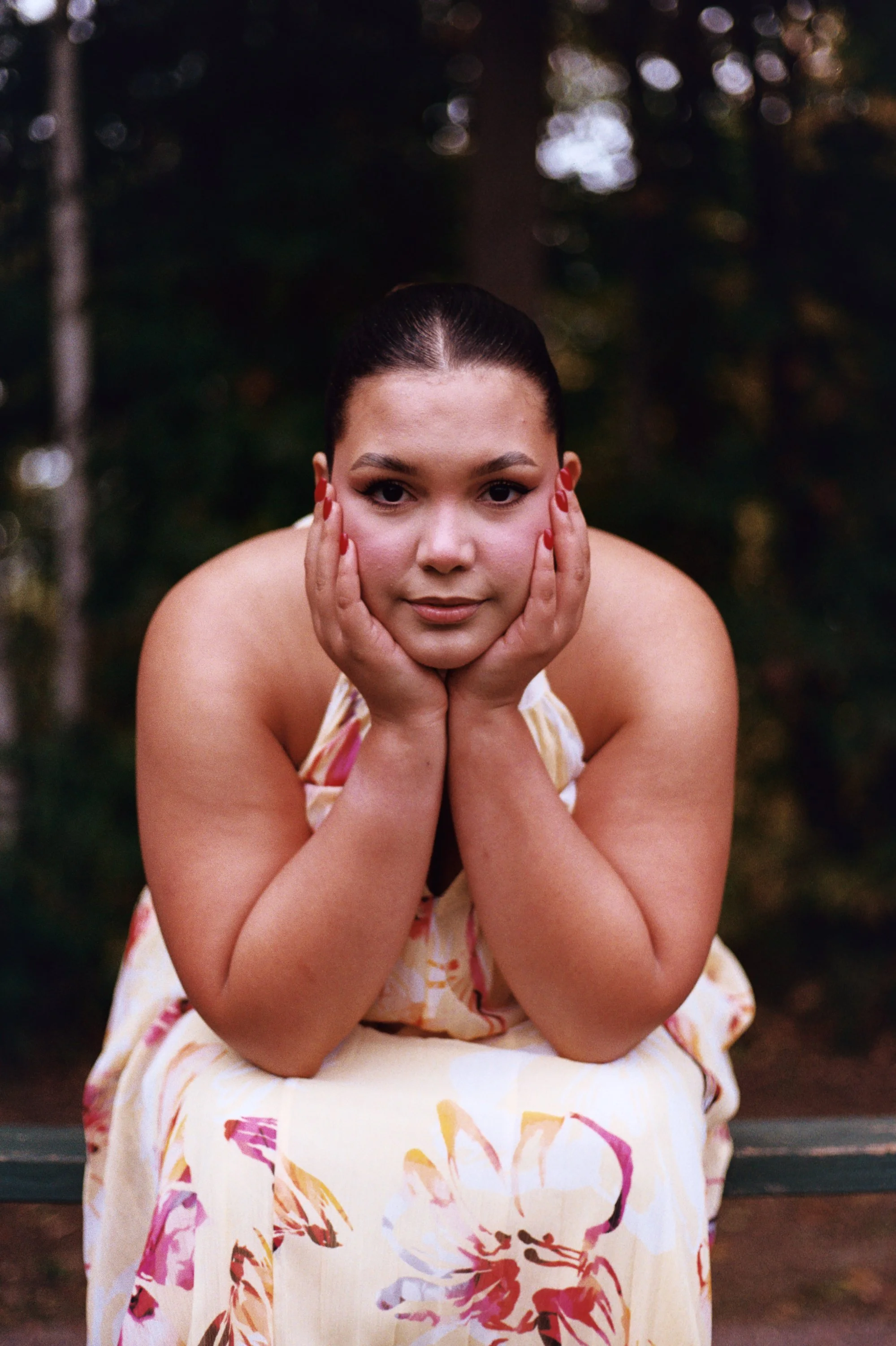 A woman with dark hair slicked back, wearing a light dress with a floral pattern, poses outdoors with her hands on her face, in front of a blurred background of trees and natural light.
