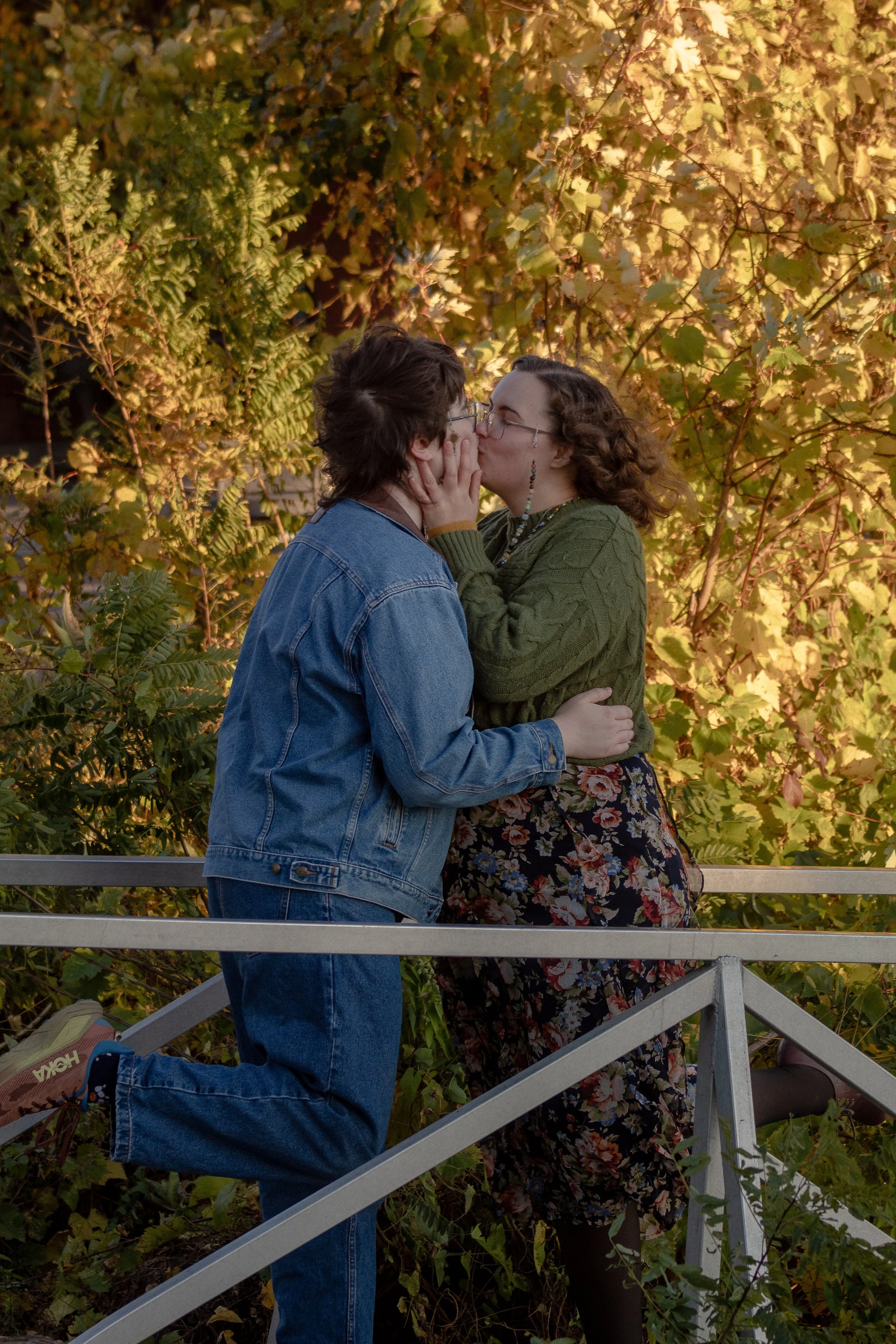 Two women are kissing on a bridge surrounded by autumn foliage. The woman on the left is wearing a denim jacket and jeans, and the woman on the right is wearing a green sweater, floral skirt, and earrings. They are holding each other close, sharing a