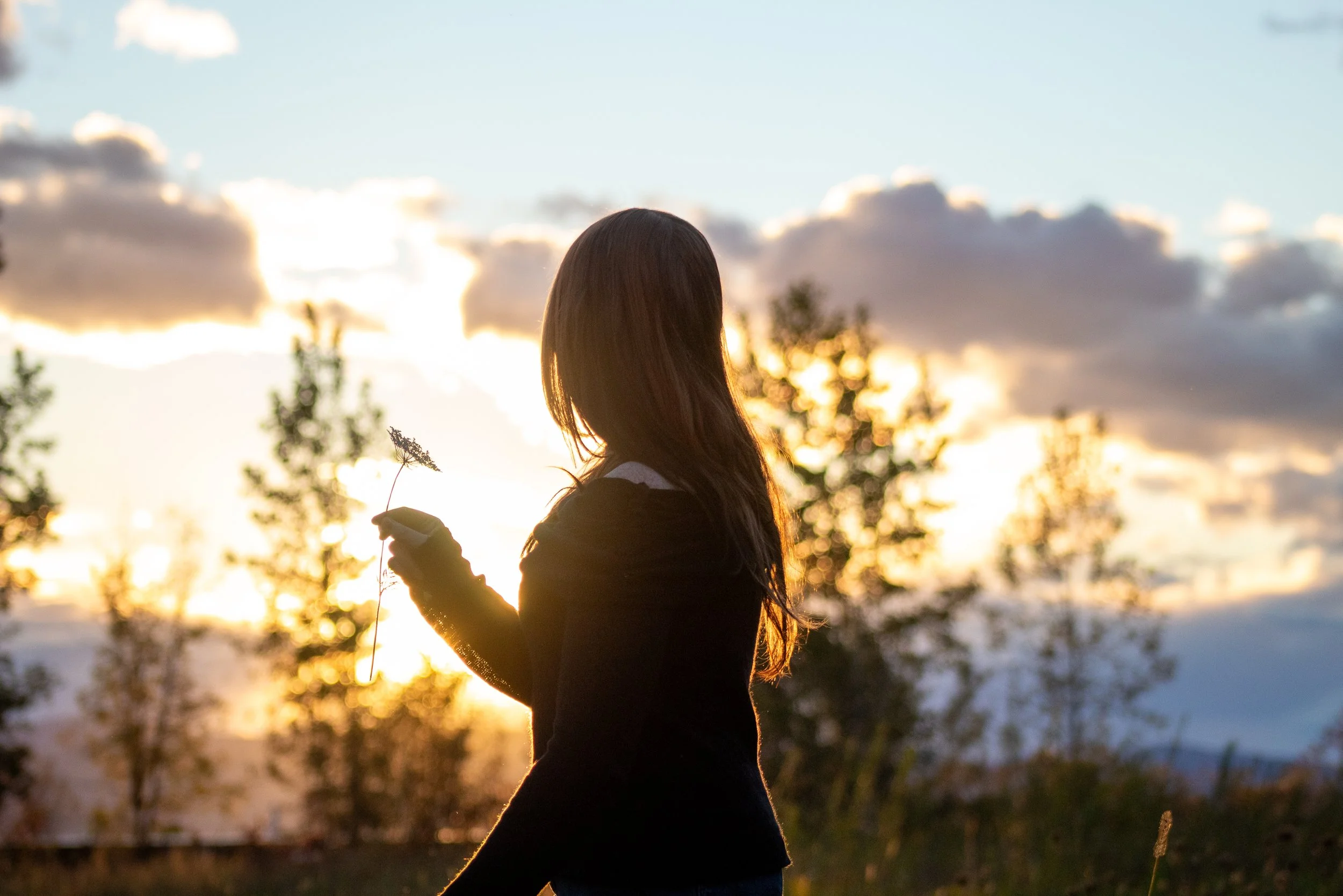 A woman standing outdoors during sunset, holding a plant and looking at the sky with clouds.