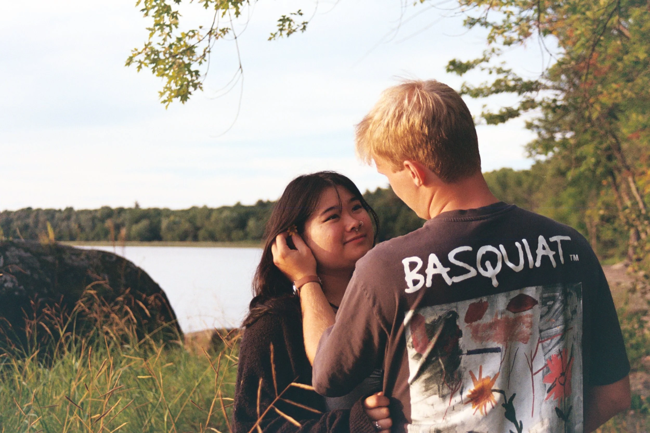 A young woman and a young man standing outdoors near a river, sharing a tender moment. The woman is looking up at the man with a smile, while the man is gently holding her face. The background features trees with green and orange leaves, indicating a