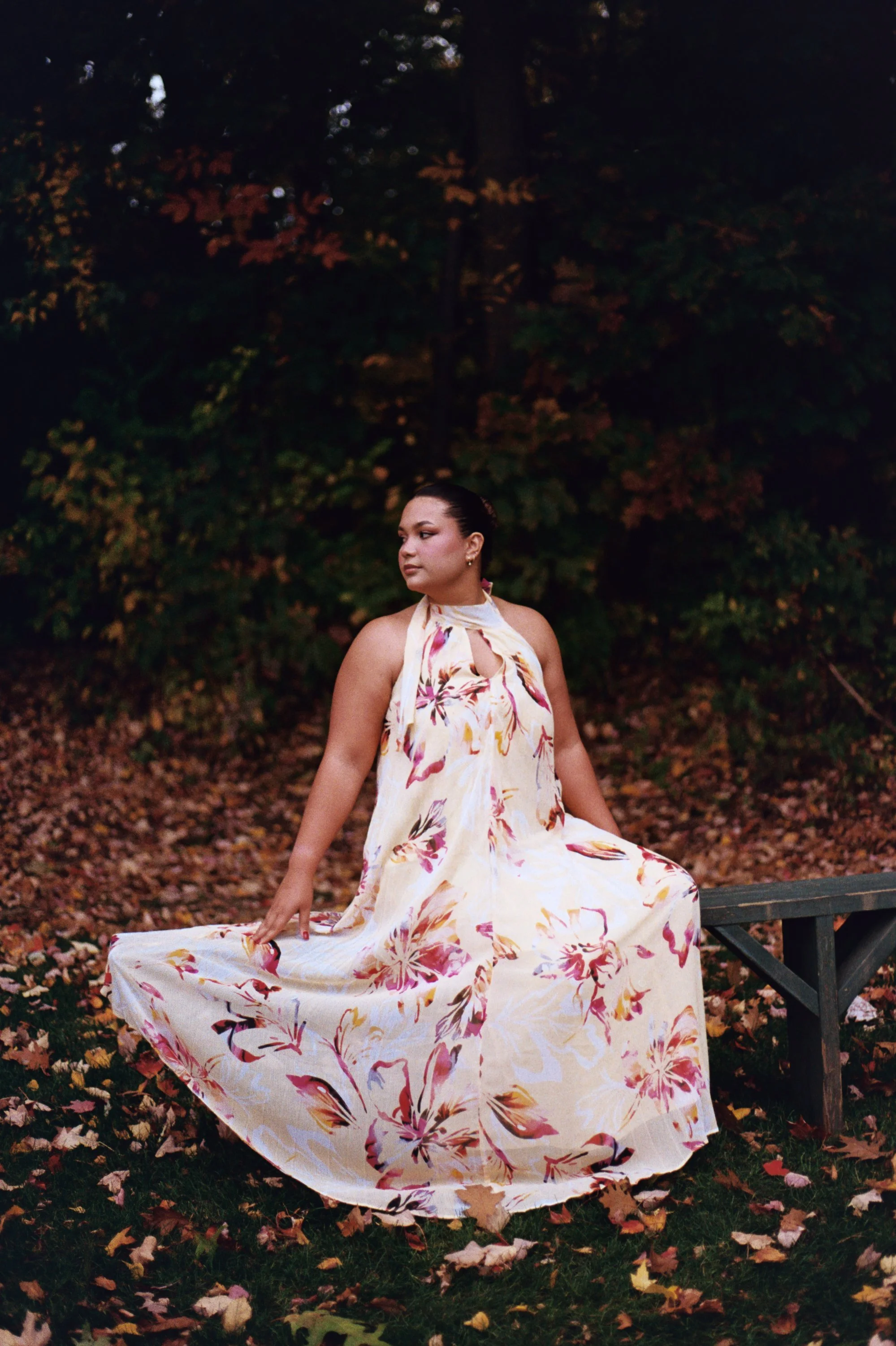 A woman in a flowing dress with pink and orange floral patterns sitting on a bench outside in a park at dusk, surrounded by fallen autumn leaves and trees with darkened foliage.