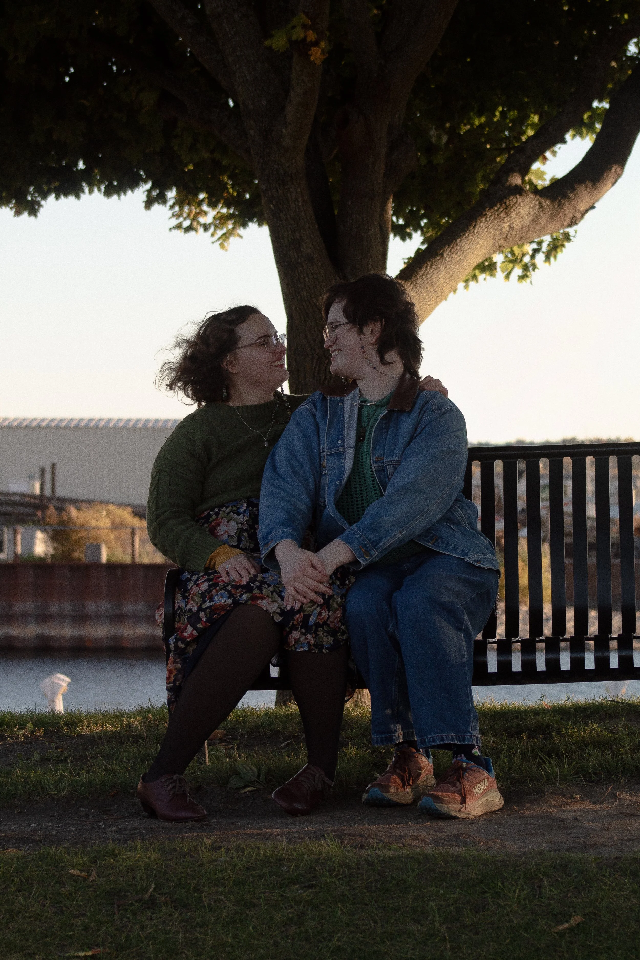 Two women sitting on a park bench under a large tree, smiling at each other, during sunset or early evening.
