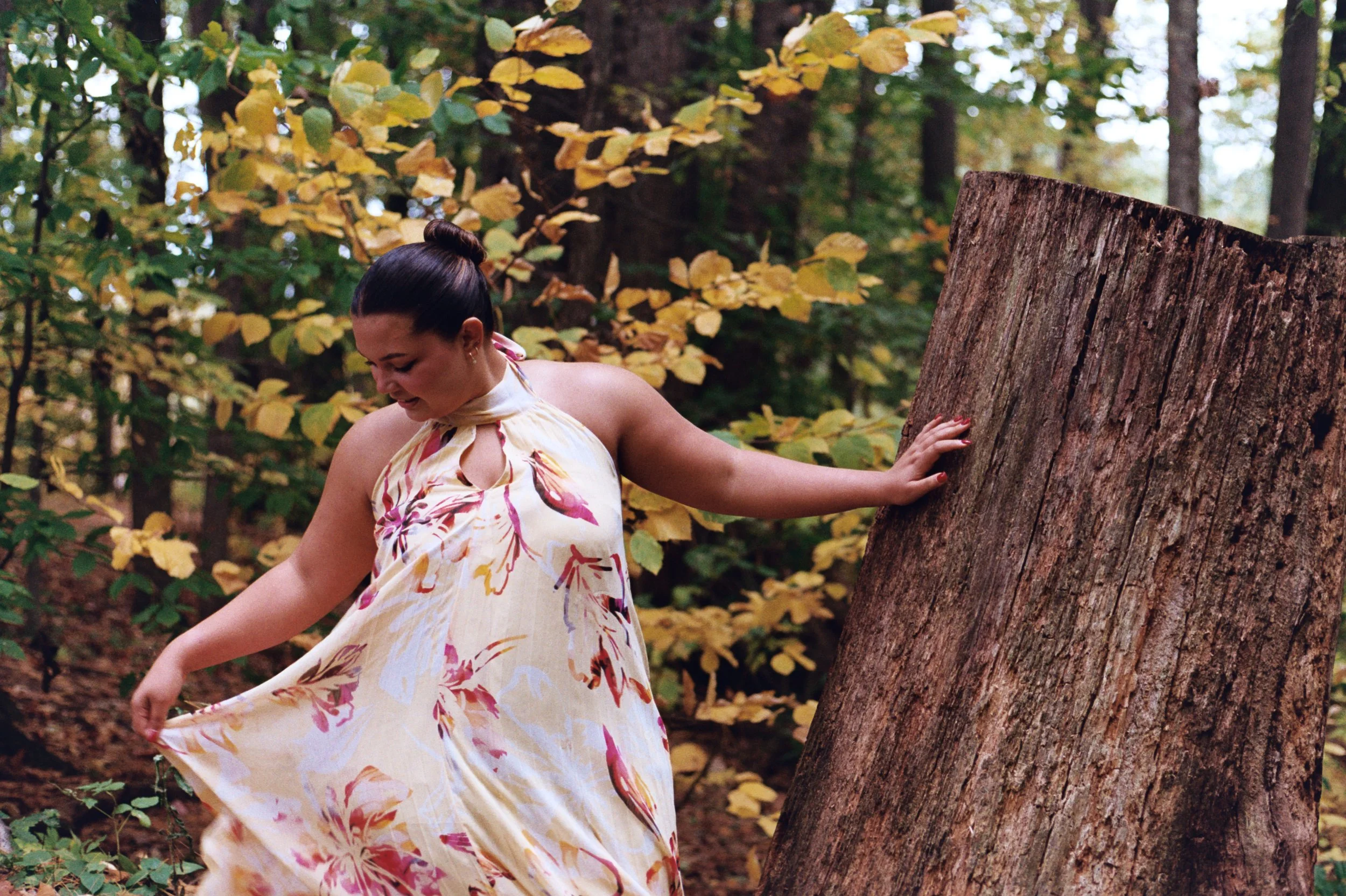 A woman with dark hair tied in a bun wears a colorful floral dress and stands in a forest, touching a large tree trunk surrounded by green and yellow foliage.