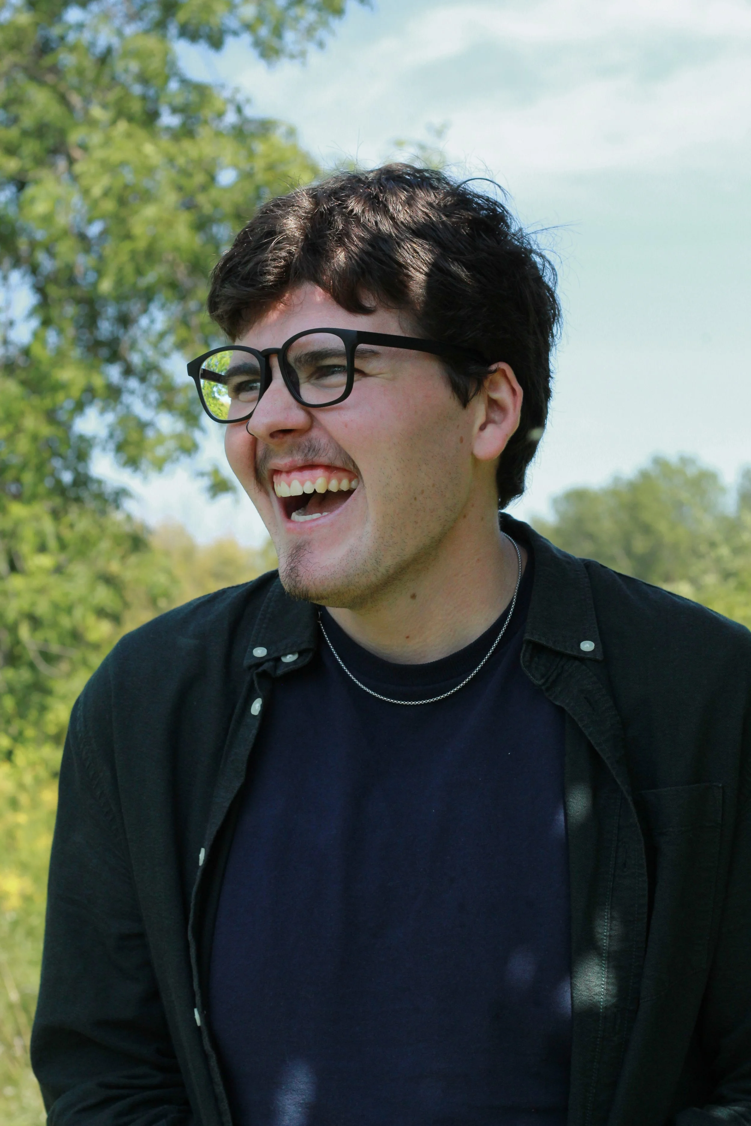 Young man smiling outdoors wearing glasses, black jacket, and navy shirt, with trees and a cloudy sky in the background.