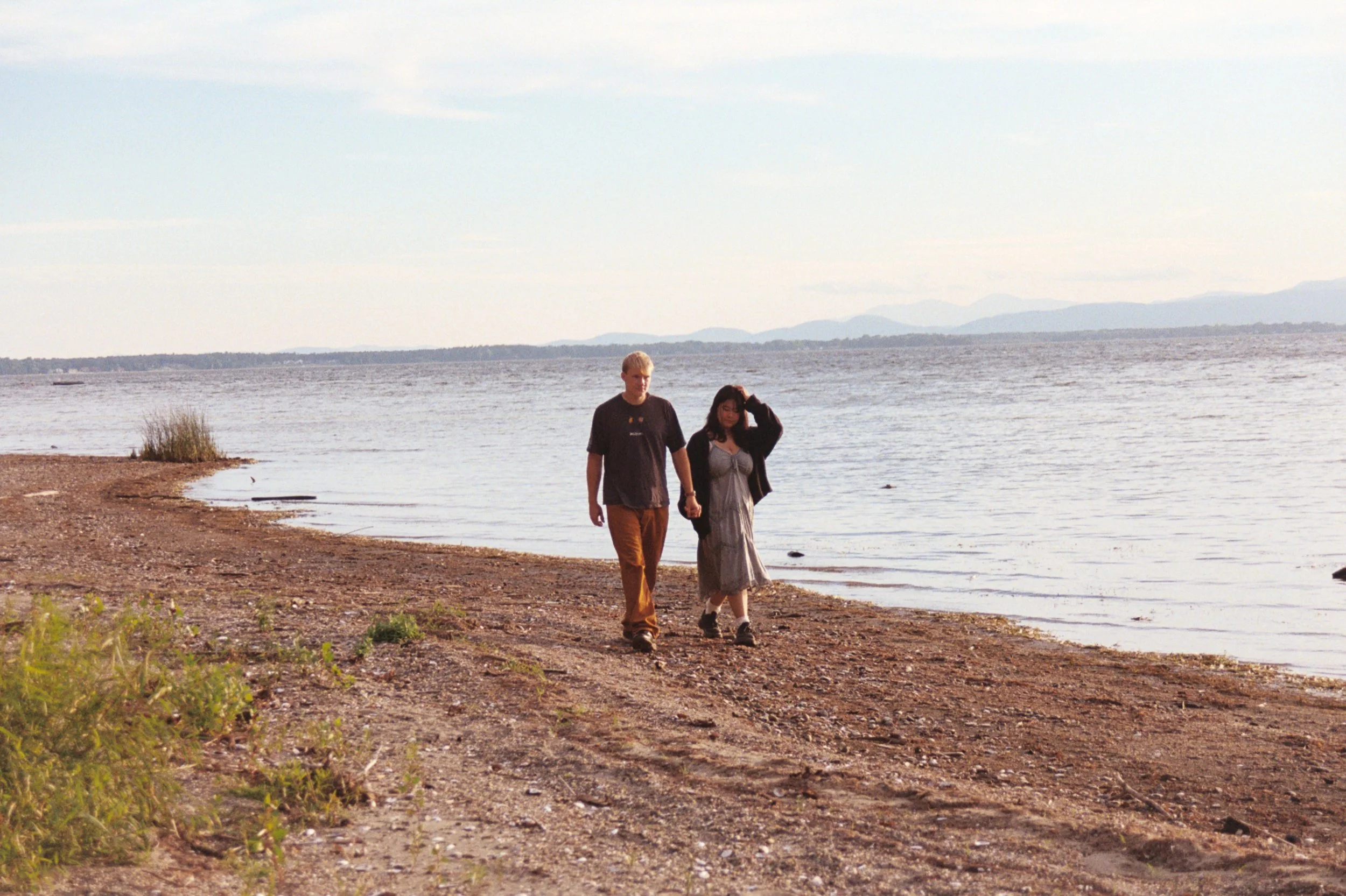 A couple walking hand in hand along a pebbled beach near a body of water, with mountains in the distance under a cloudy sky.