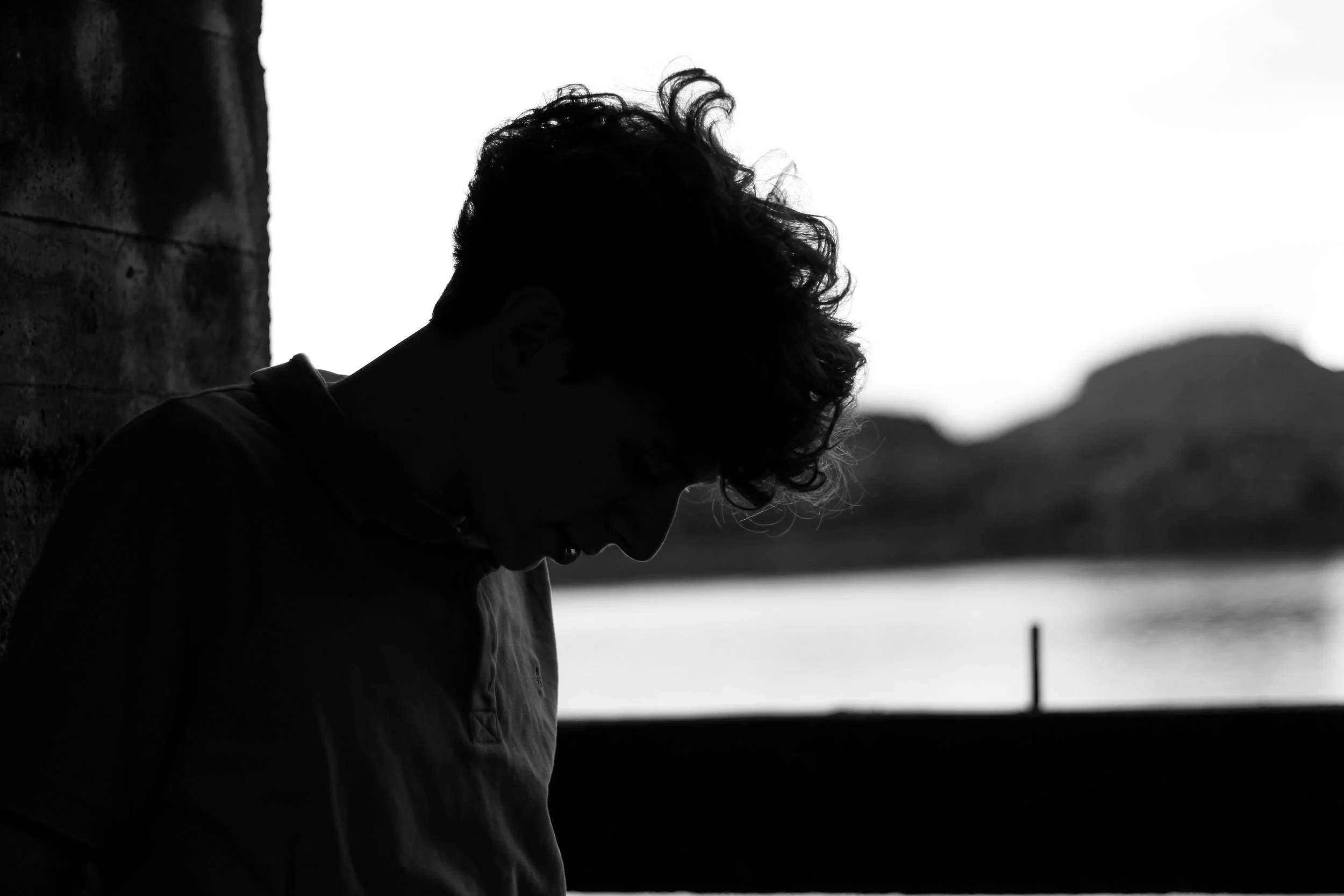 A black and white silhouette of a person with curly hair, looking down while standing near a stone wall with water and mountains in the background.