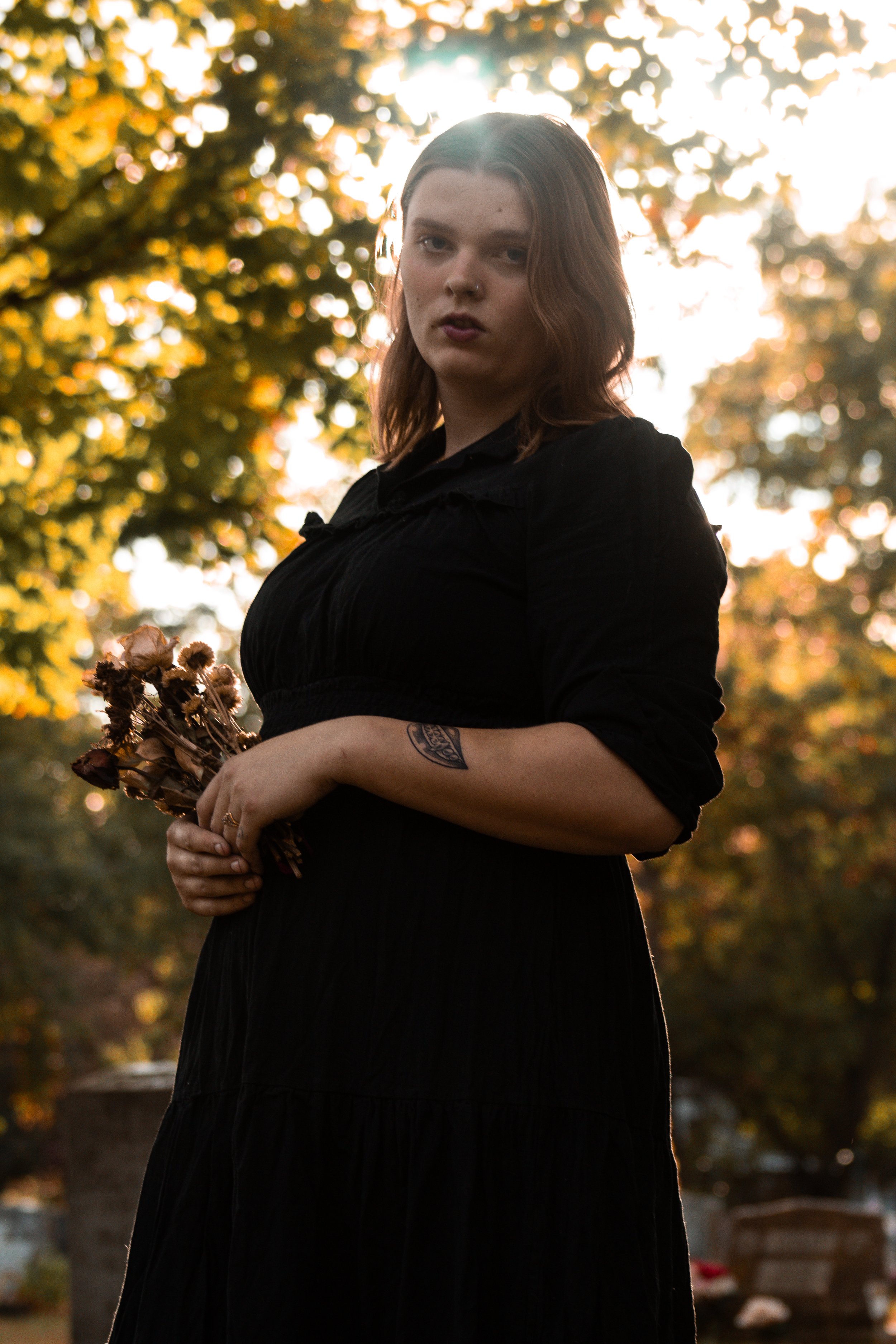 A young woman with long brown hair and a septum piercing wearing a black dress, standing outdoors at sunset, holding a bunch of dried flowers, with trees and a cemetery in the background.