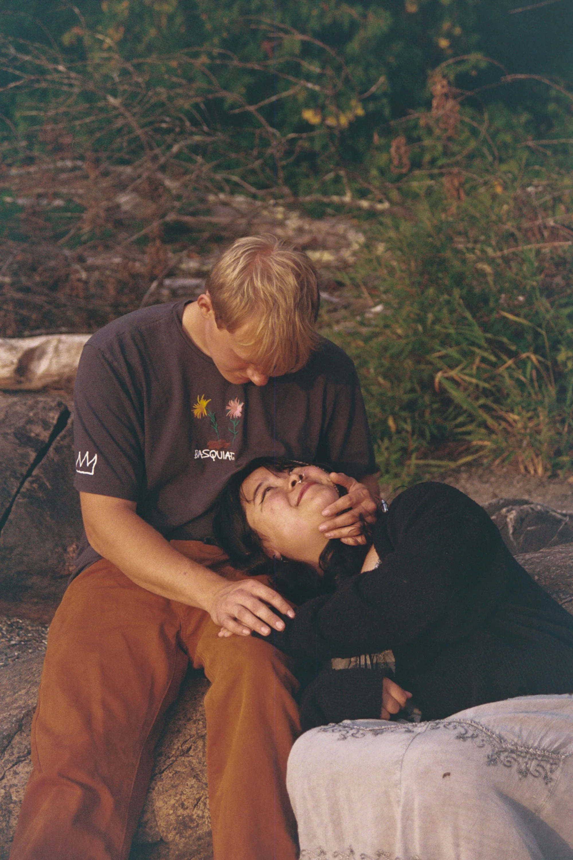 Two people, one lying on the ground and the other kneeling beside, appear to be performing CPR or rescue procedures outdoors at night, with branches and foliage in the background.