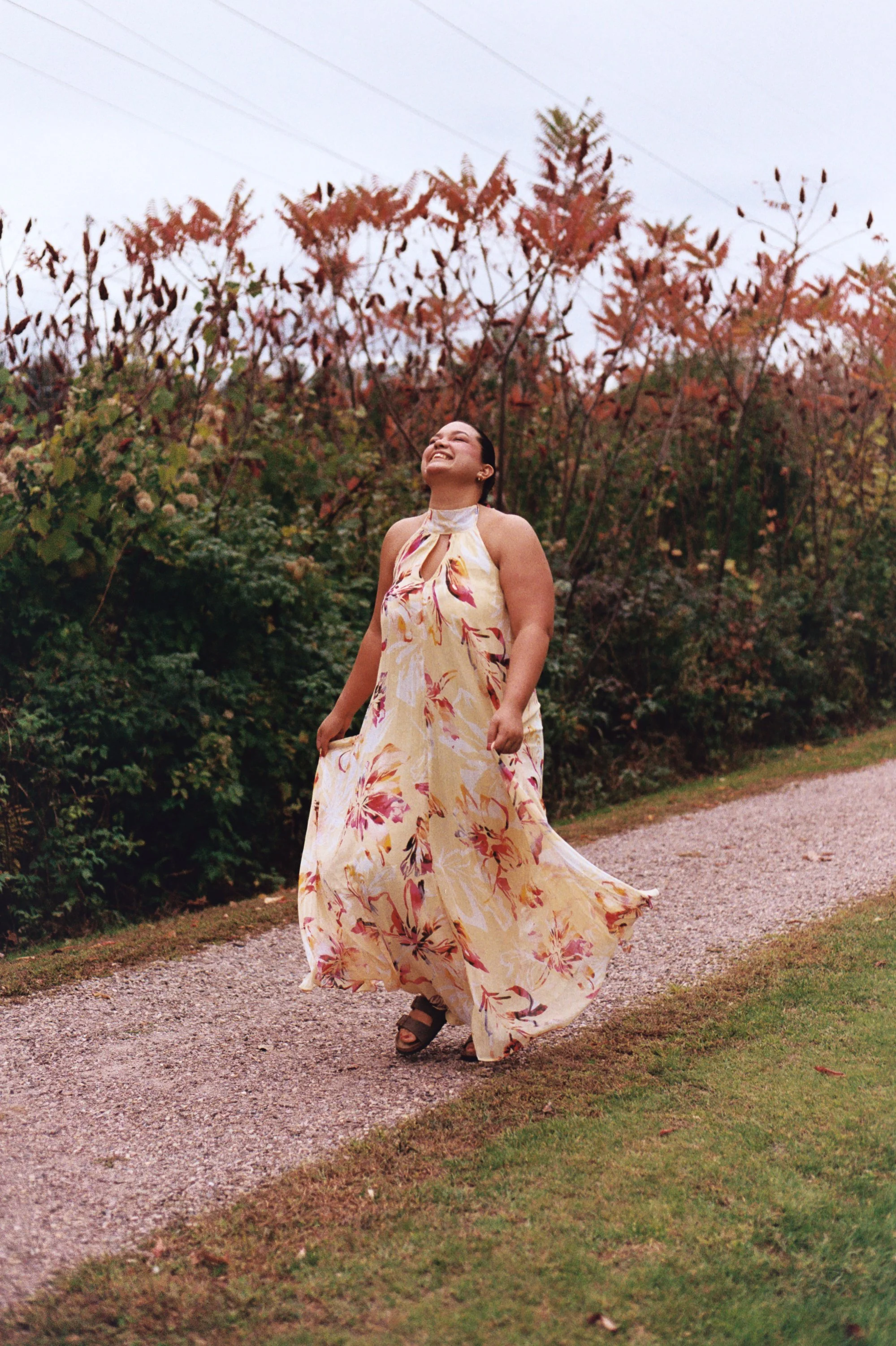 A woman with dark hair tied back, smiling and looking upward, walking along a gravel path surrounded by lush greenery and trees with autumn-colored leaves, wearing a flowy yellow floral dress and sandals.