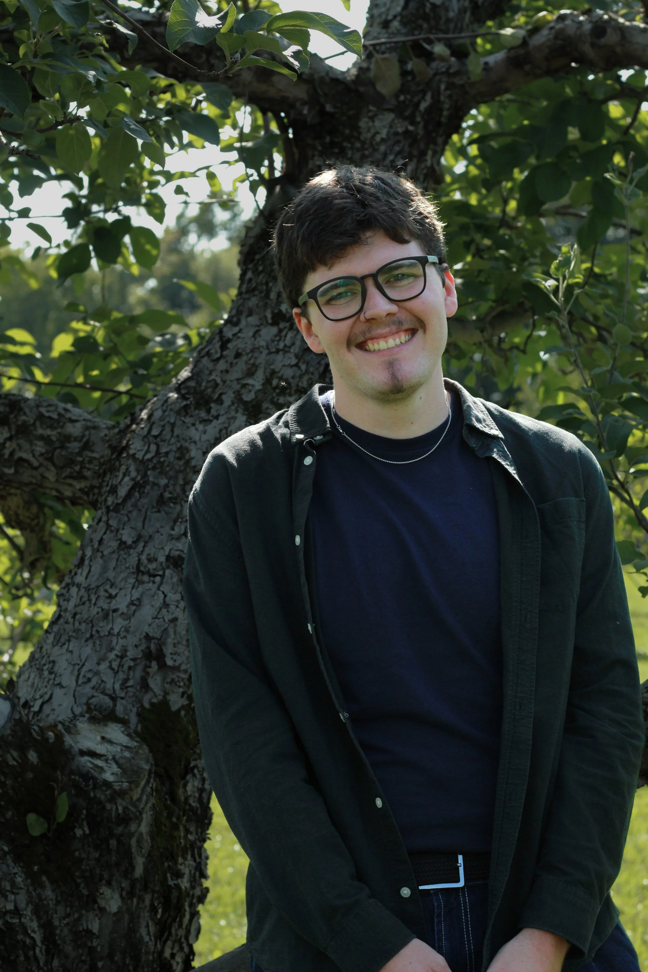 A young man with glasses and dark hair smiling outdoors, standing in front of a tree with green leaves.