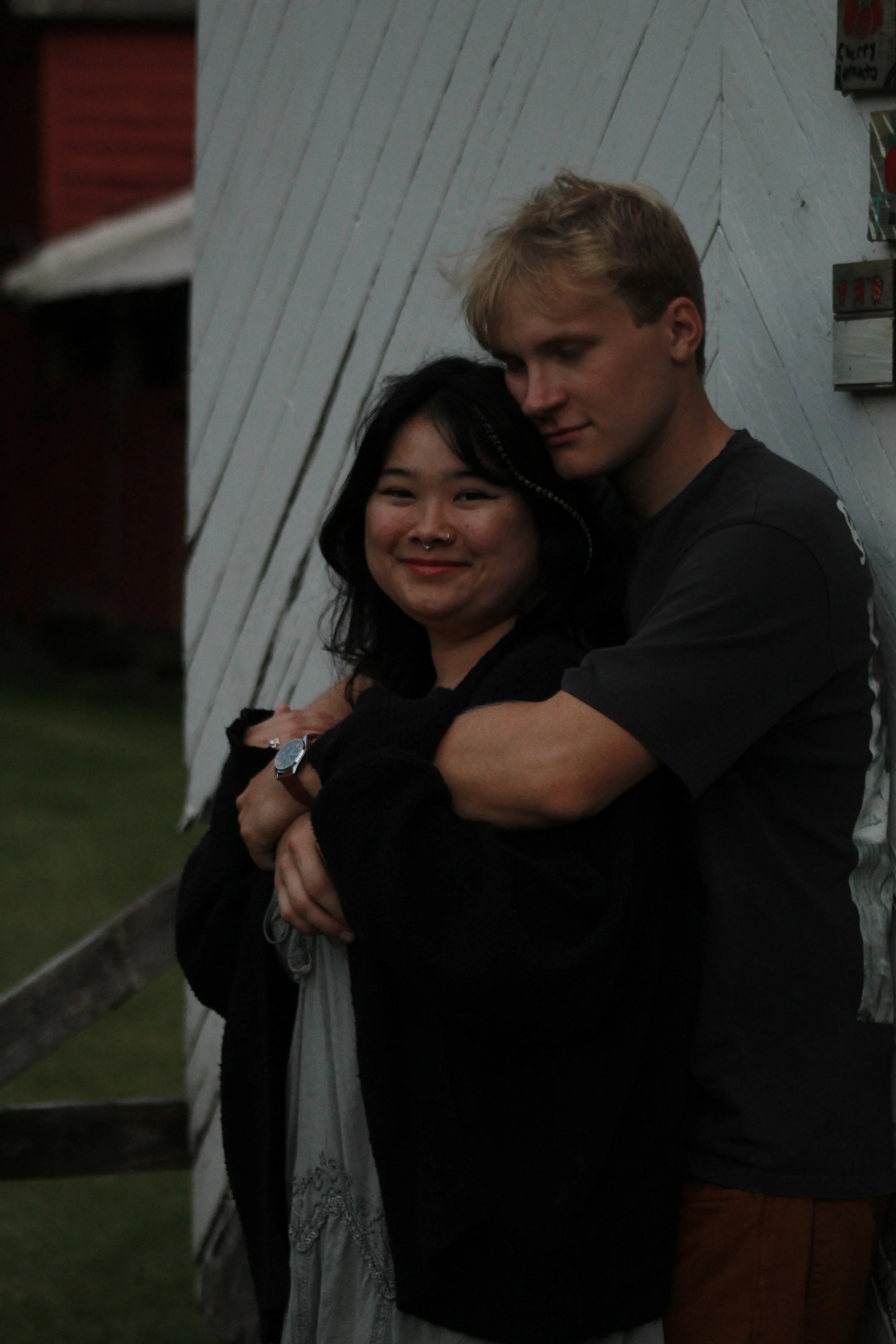 A young woman with dark hair and a nose piercing, and a young man with blonde hair, hugging each other affectionately against a white wooden-paneled wall, outdoors during the evening.