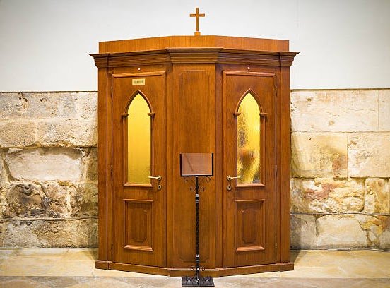 A wooden confession booth with two doors featuring arched windows, set against a stone wall inside a church, with a small stand and microphone in front.