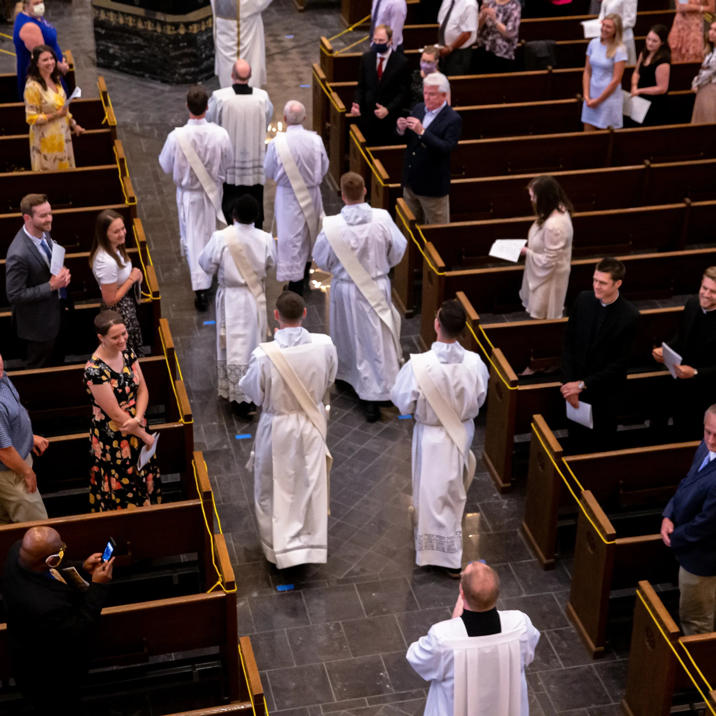 People attending a church service with some wearing masks. A priest is standing at the altar with clergy members, while others are standing and seated in the pews, some holding papers or forming an orderly arrangement.