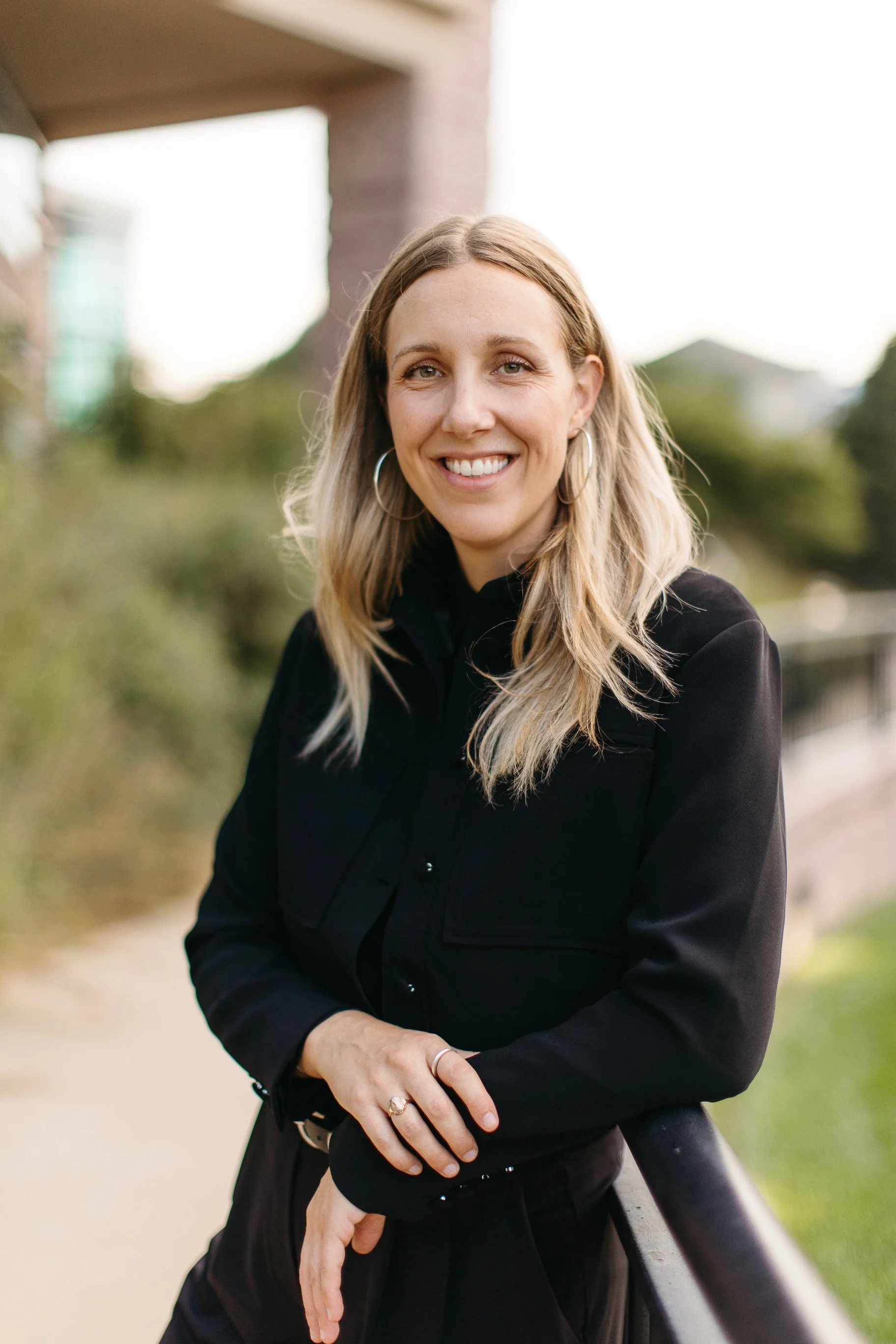 A woman with blonde hair and hoop earrings smiling outdoors, wearing a black jacket, with greenery and a pathway in the background.
