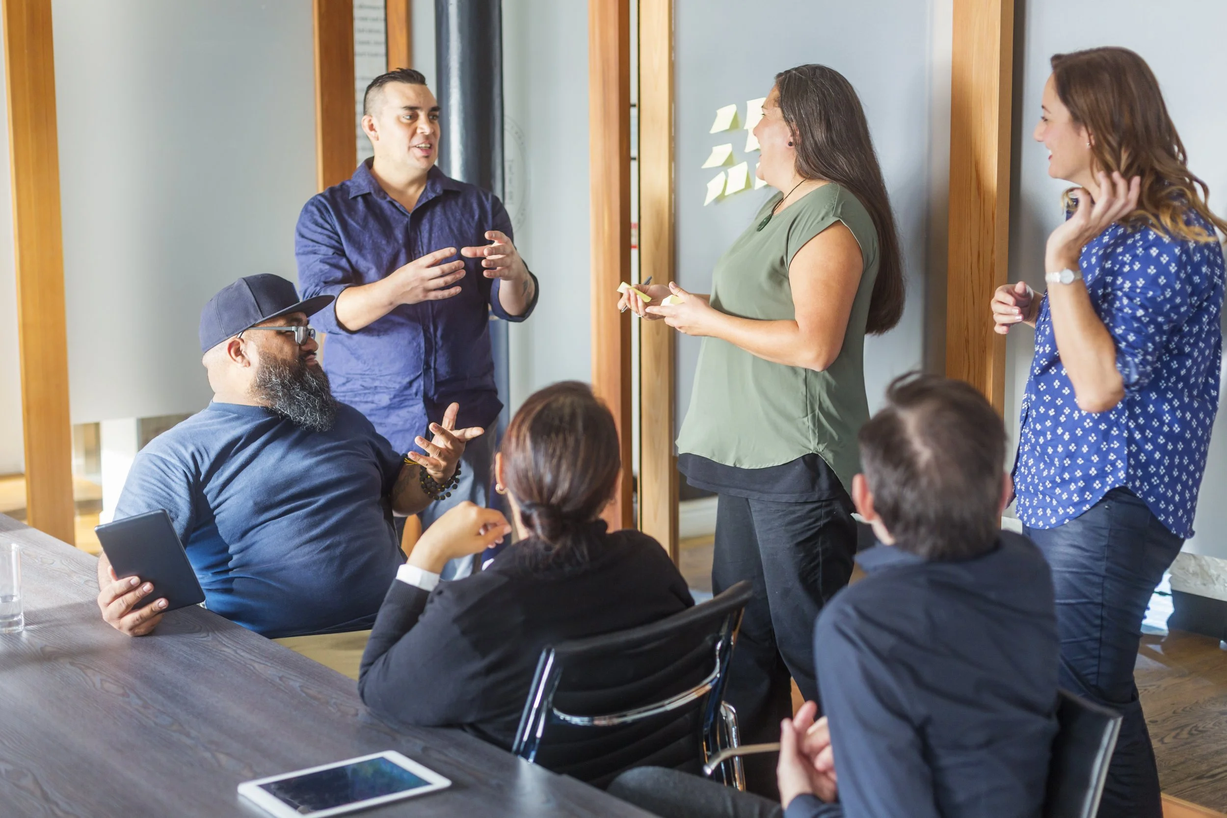 A group of five people engaged in a discussion in a professional setting, with some people sitting at a table and others standing near a wall with sticky notes.