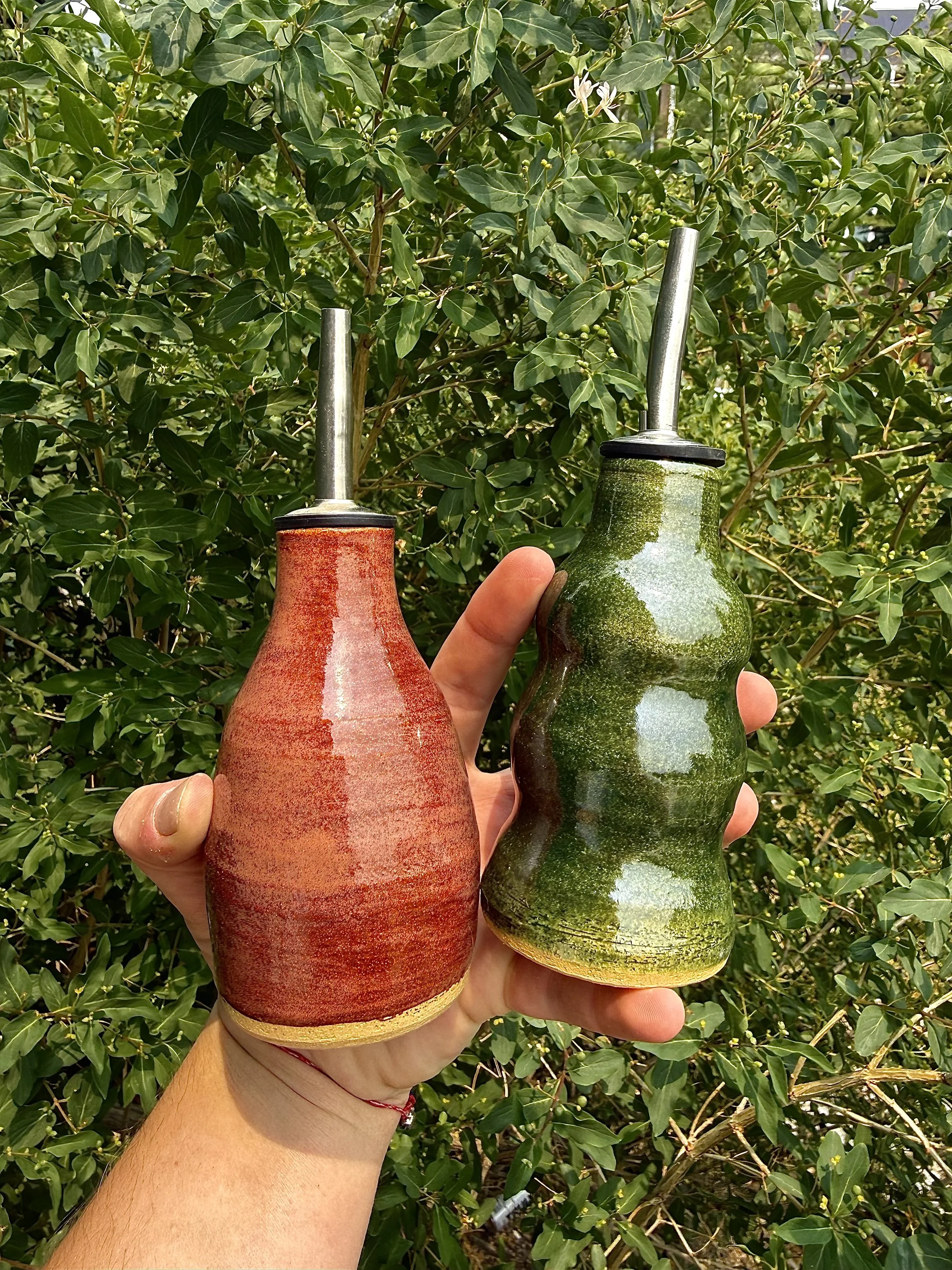 A person holding two ceramic watering cans in front of a green leafy bush. One watering can is red and the other is green.