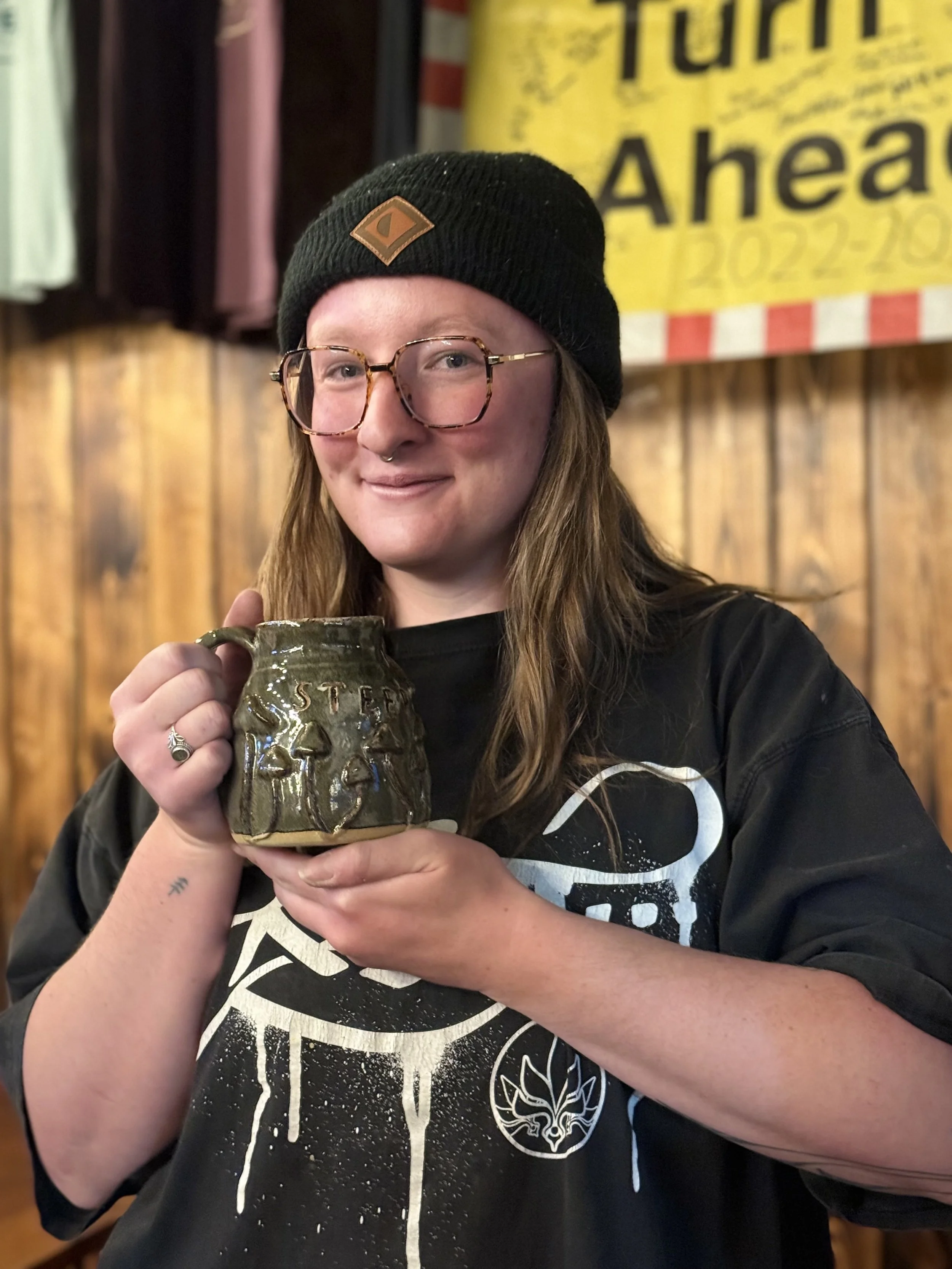 A woman with long brown hair, glasses, and a beanie hat holding a ceramic mug with the word 'STEEL' on it, smiling in a wooden interior with a banner in the background.