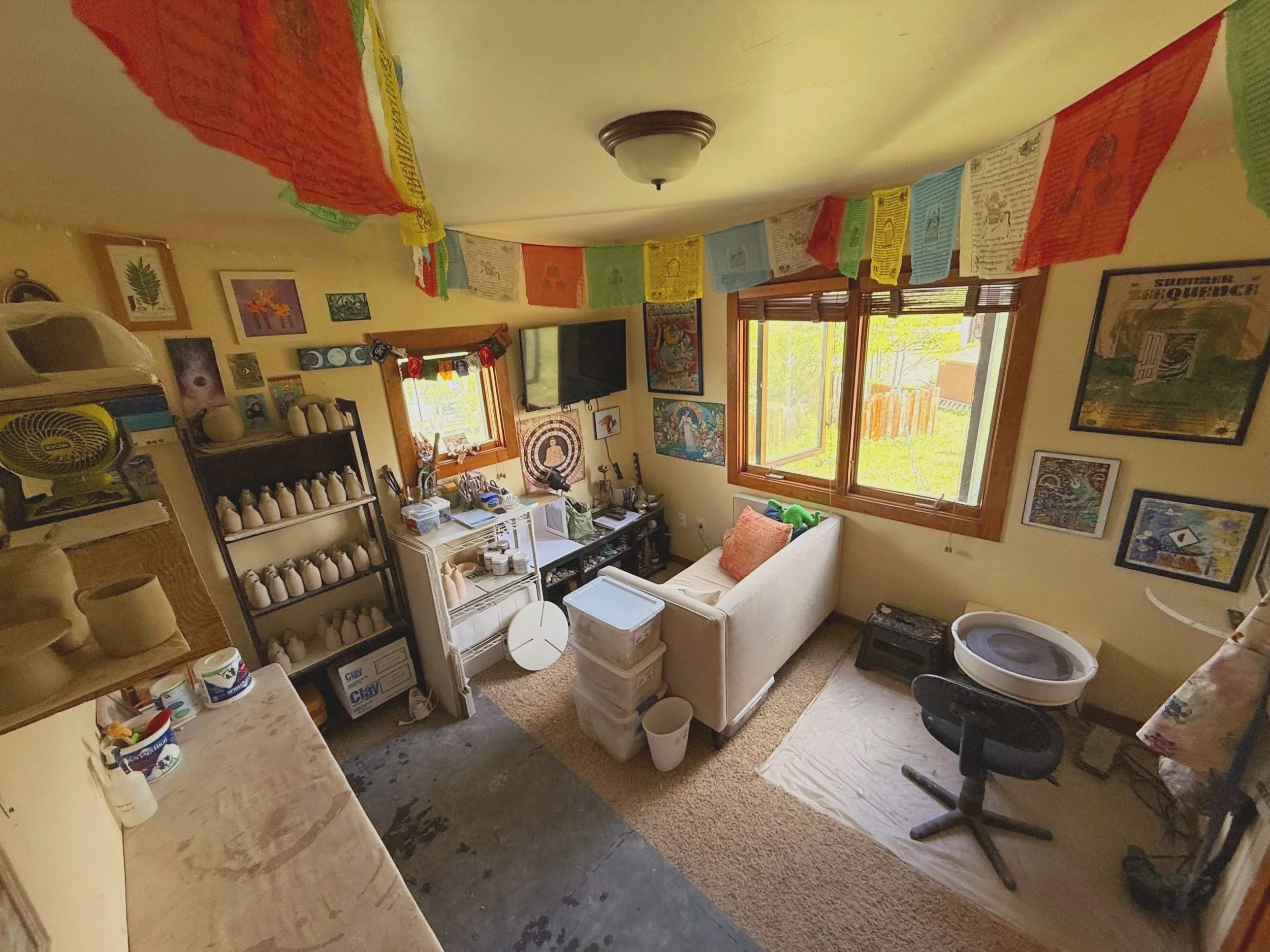 A cluttered living room filled with colorful prayer flags hanging from the ceiling, various artworks on the walls, a sofa with a throw pillow, a black office chair, multiple storage containers, and miscellaneous household items, with windows showing a view of trees outside.