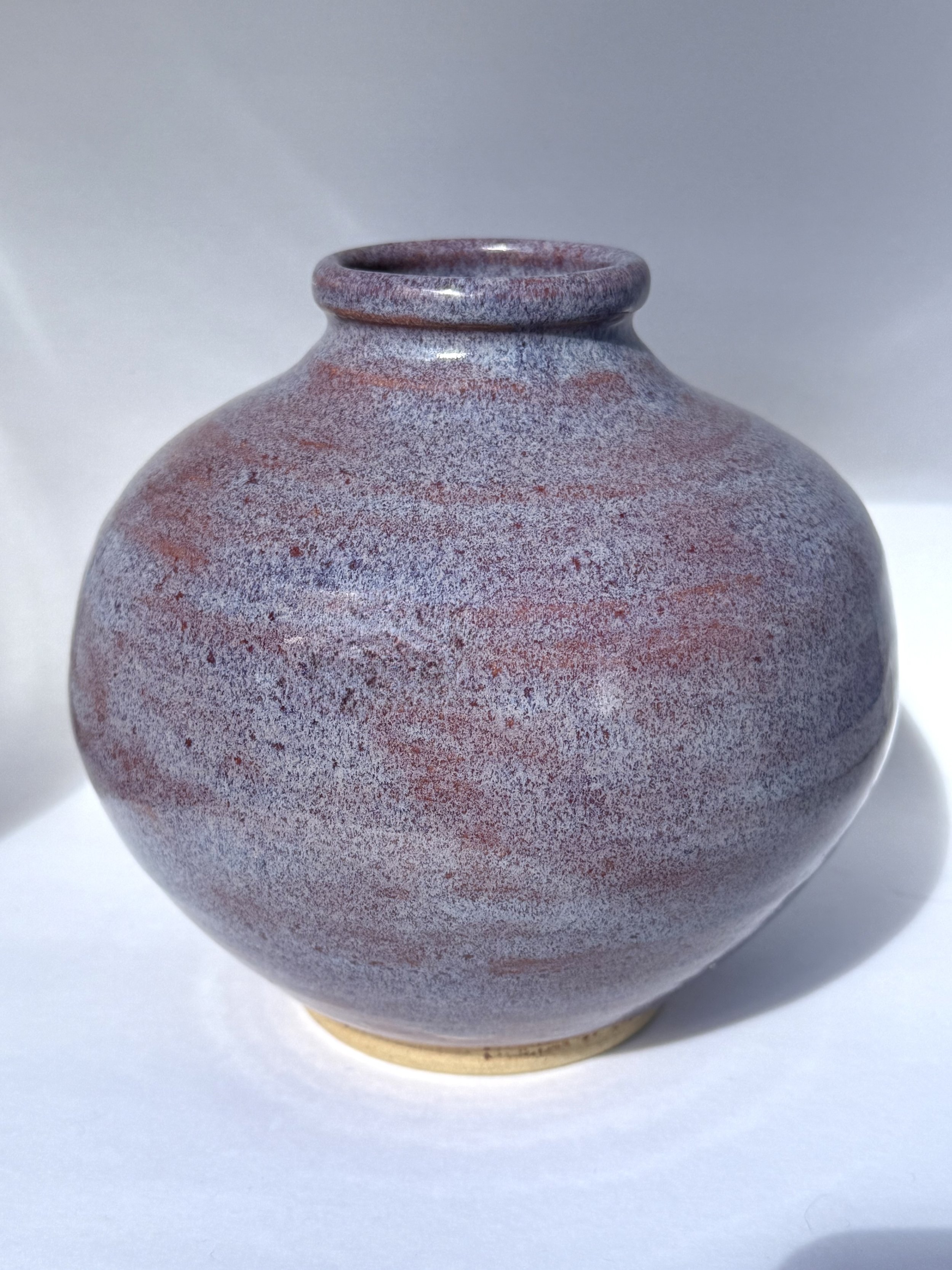 A ceramic vase with a round body and narrow neck, glazed in shades of purple, red, and gray with a speckled appearance, set against a plain white background.